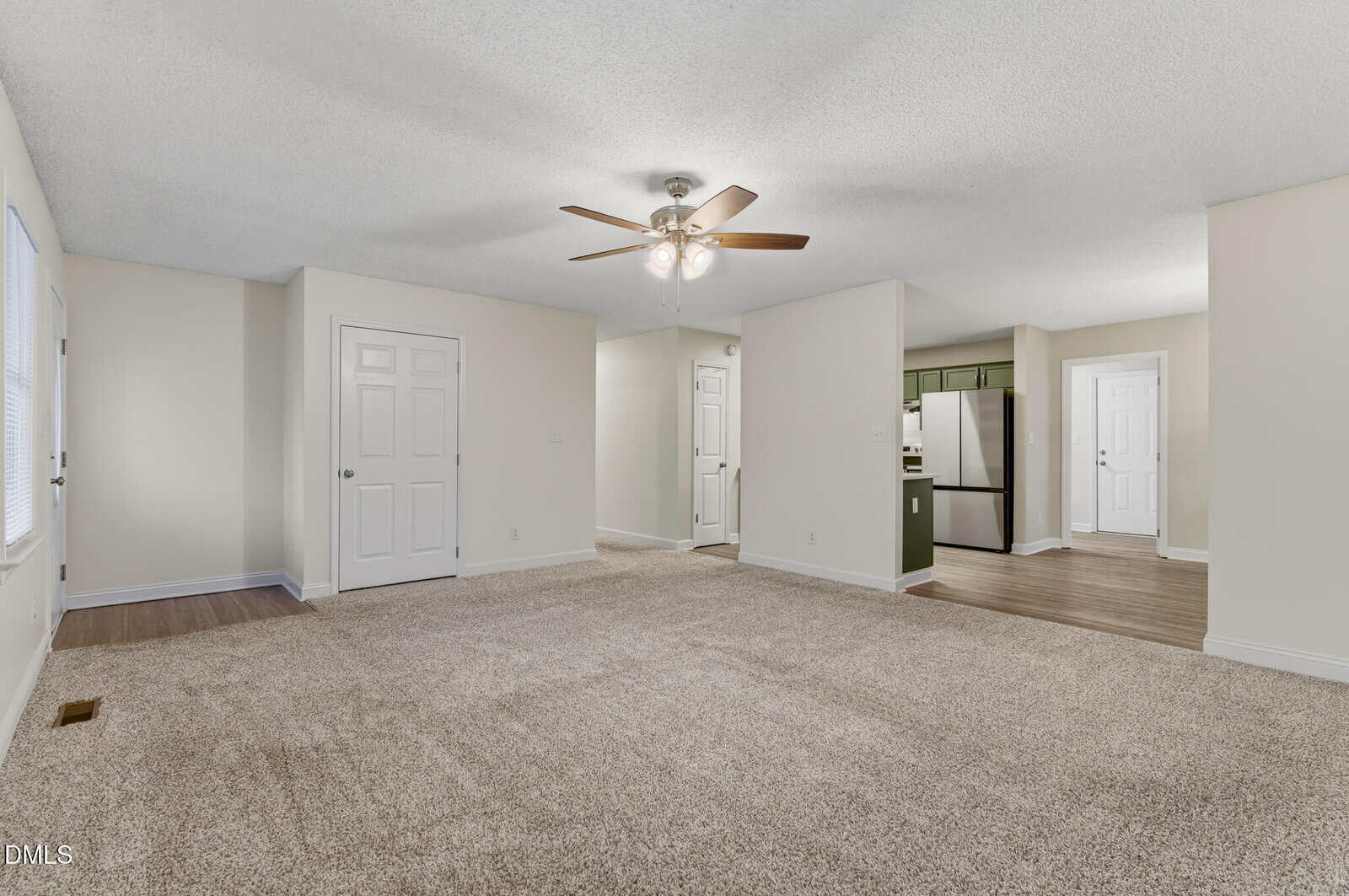 Spacious living area with beige carpet, ceiling fan, and white walls. The room opens to a kitchen with stainless steel appliances and green cabinets, and multiple white doors are visible.