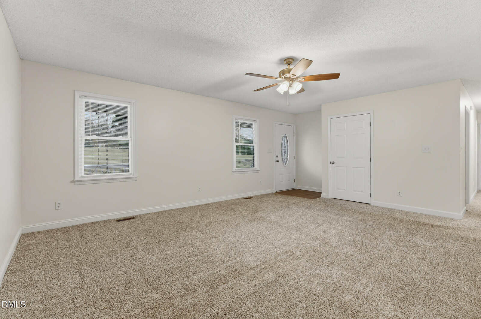 A spacious living room with beige carpet, cream-colored walls, ceiling fan with lights, two windows, a white front door with a glass oval insert, and a white interior door.