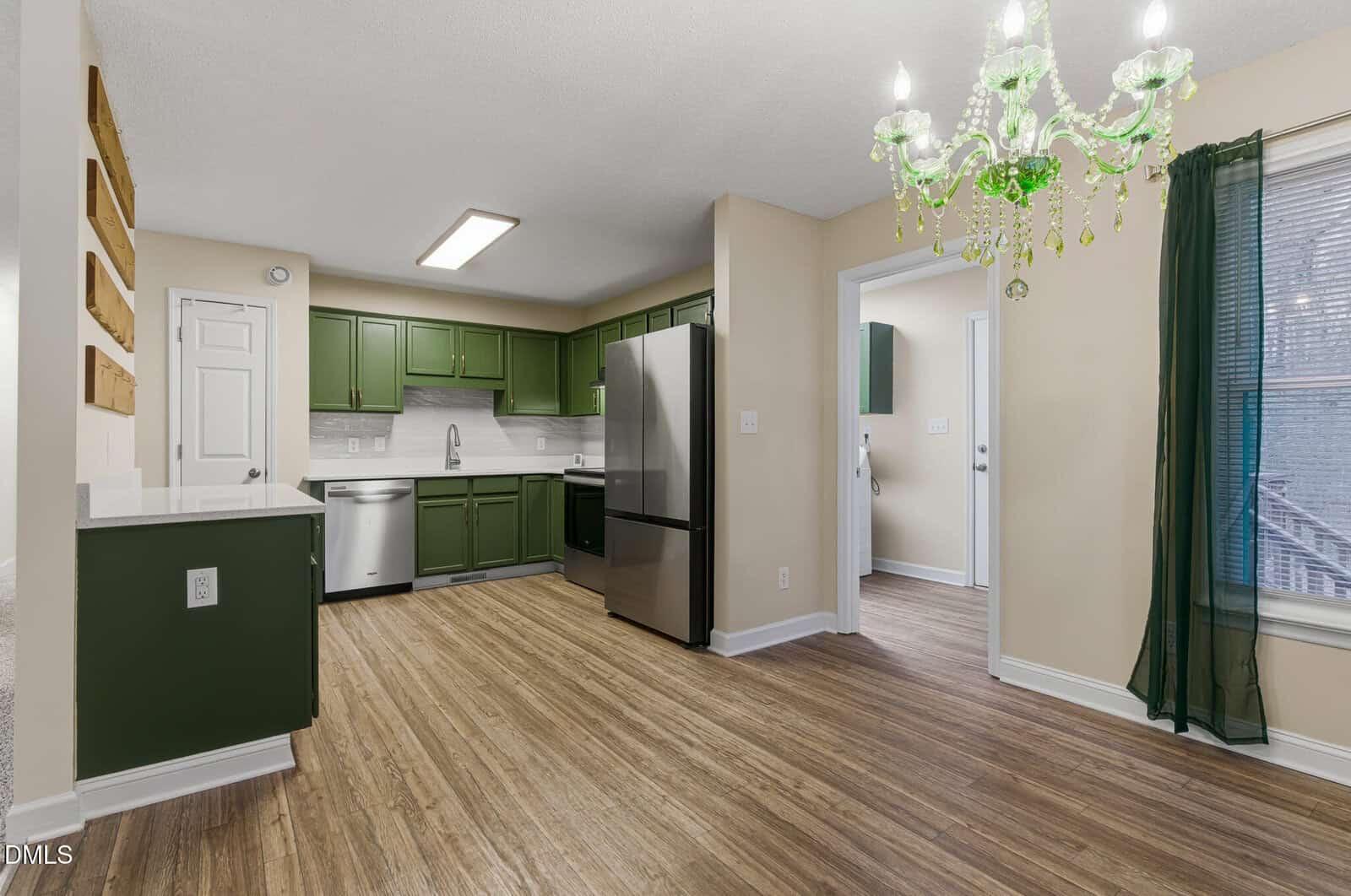 A modern kitchen with green cabinets, stainless steel appliances, wood flooring, a green chandelier, and a large window with a sheer green curtain letting in natural light.