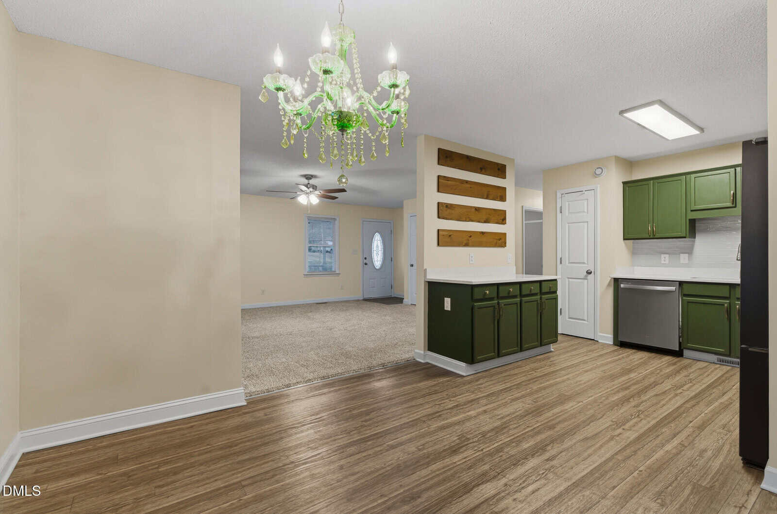 Open kitchen and dining area with green cabinets, wood flooring, beige walls, and a green chandelier. Living room with carpet and ceiling fan visible in the background. White front door and natural light.