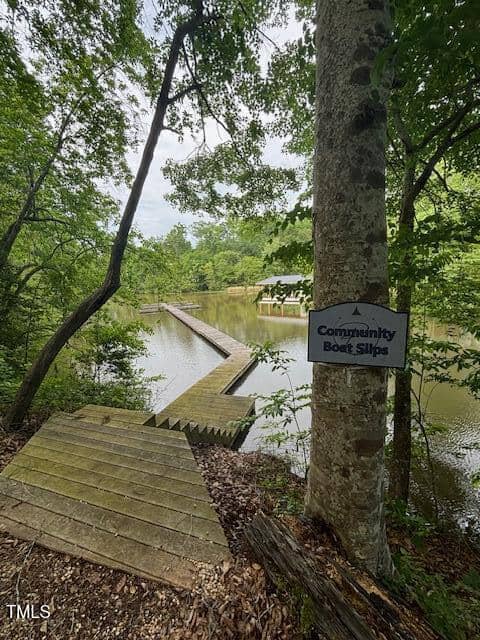 A wooden dock extends into a calm lake surrounded by trees. Steps lead down to the dock, and a sign on a tree reads “Community Boat Slips.” A covered dock structure is visible in the background.