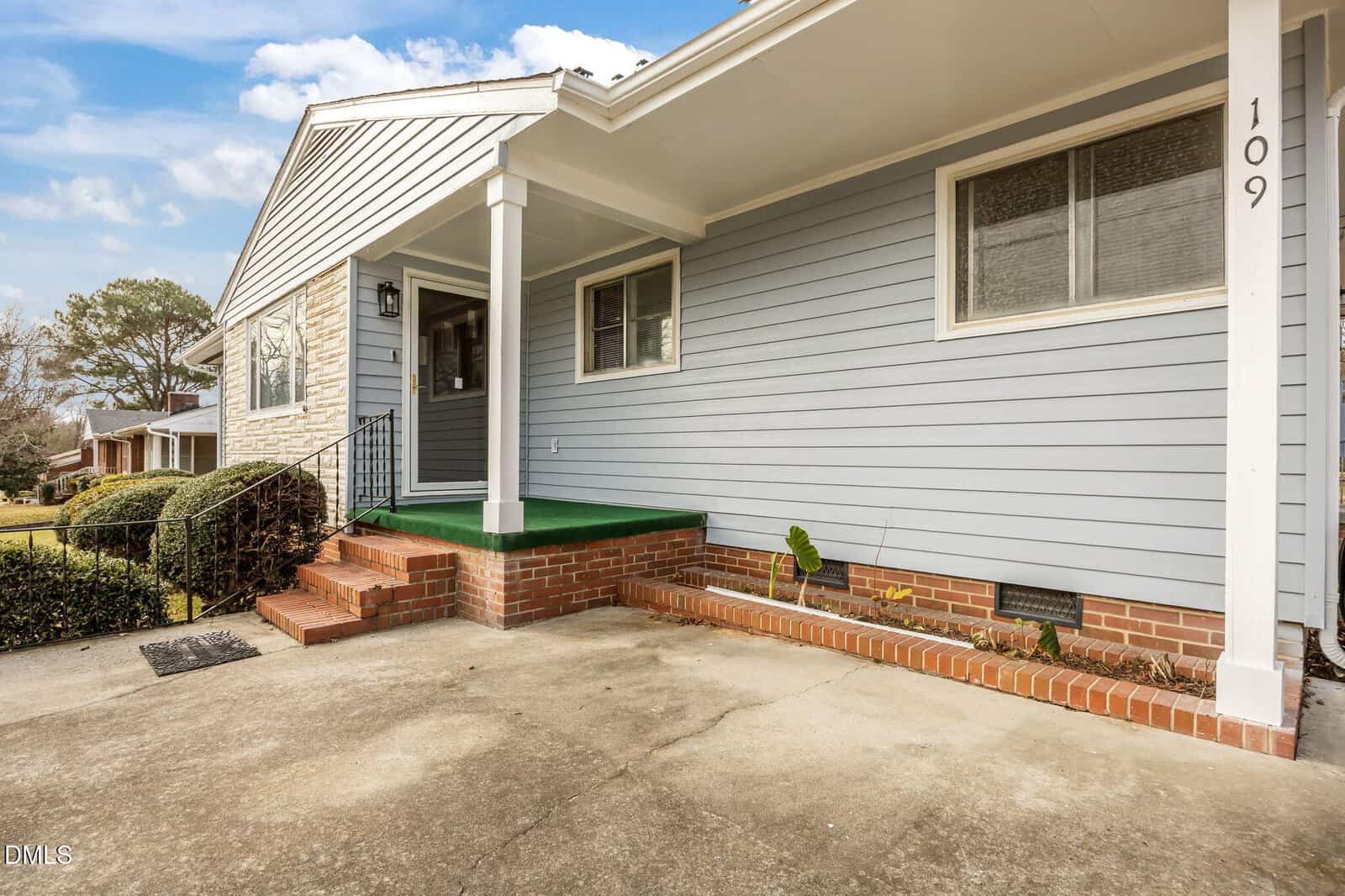 A single-story house with light blue siding, a covered front porch with green flooring, red brick steps, and a white pillar. The house number 109 is visible near a window. There is a concrete driveway in front.