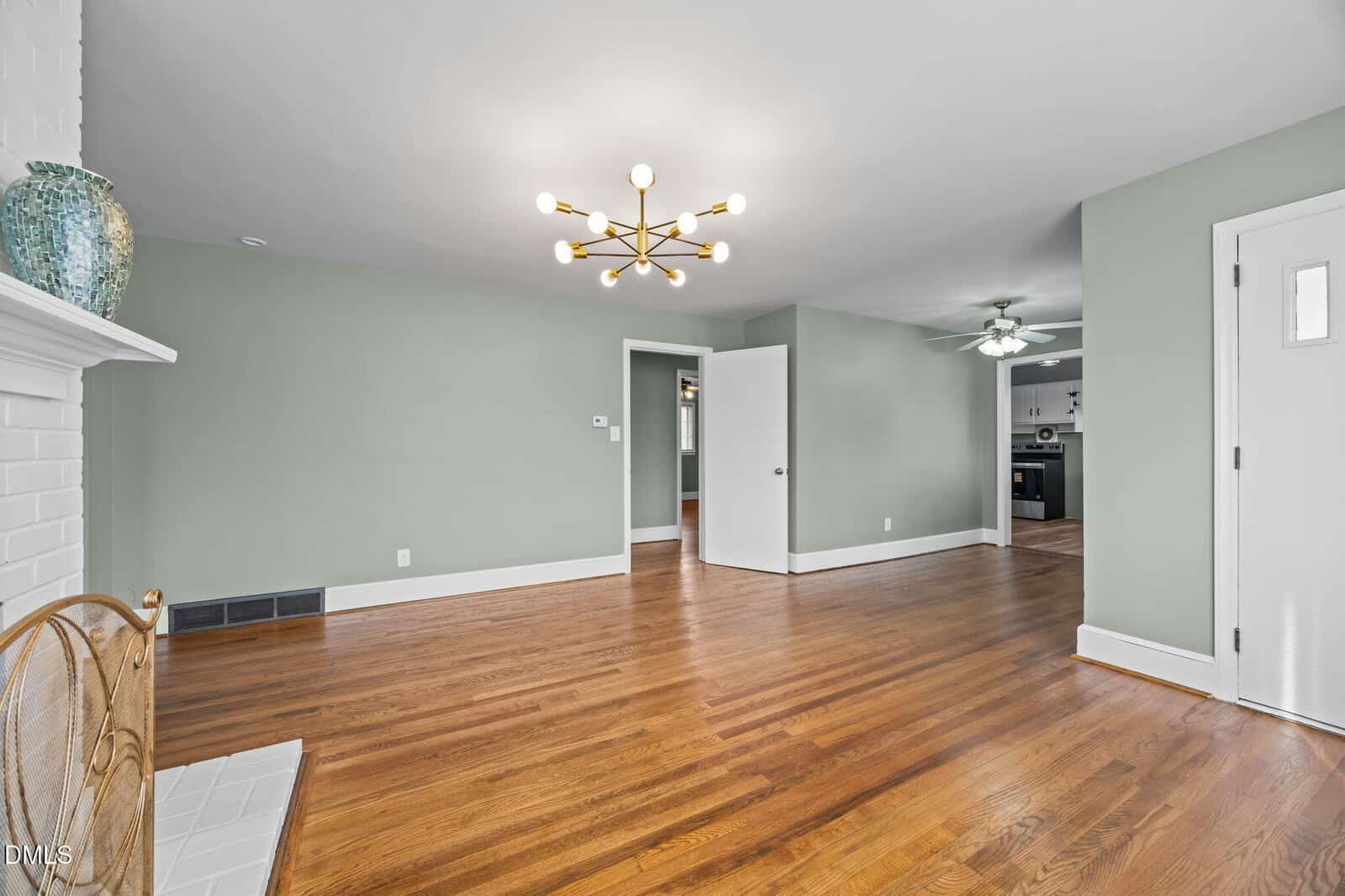 A spacious, empty living room with light green walls, hardwood floors, a modern chandelier, and a white brick fireplace. An open doorway leads to a kitchen with dark cabinets and a ceiling fan.
