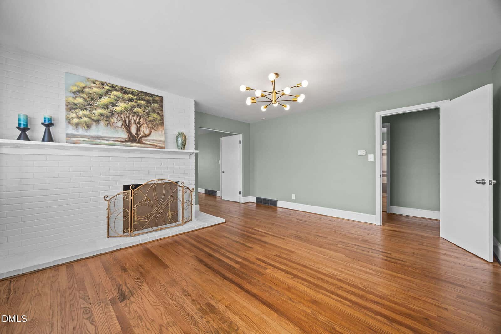A spacious living room with hardwood floors, a white brick fireplace, green walls, a modern gold chandelier, and a painting and candles on the mantel. The room has an open doorway and a partially open white door.
