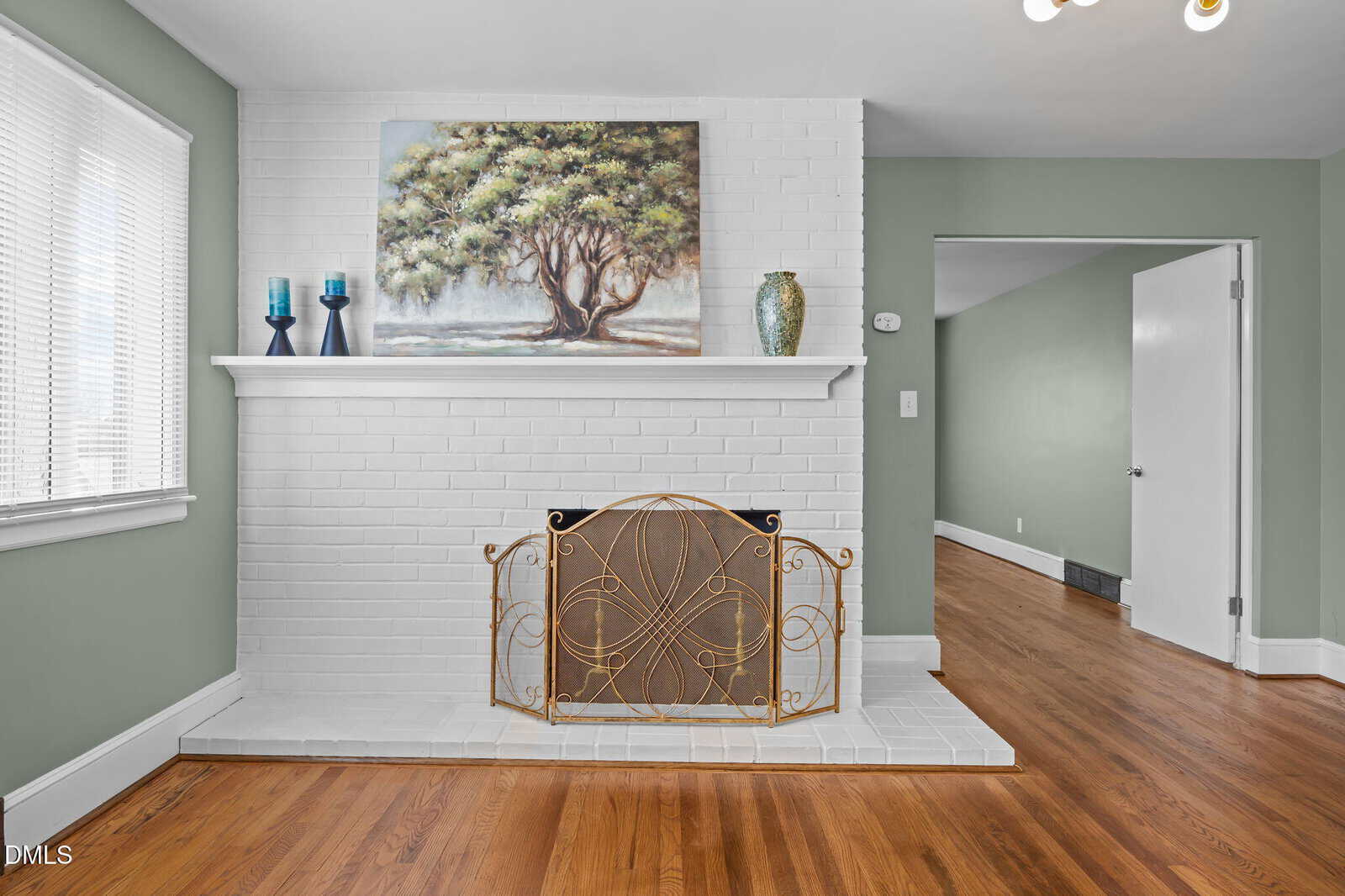 A white brick fireplace with a decorative gold screen, a mantel holding three vases and a painting of a tree. The room has light green walls and hardwood floors. An open doorway leads to an adjacent room.