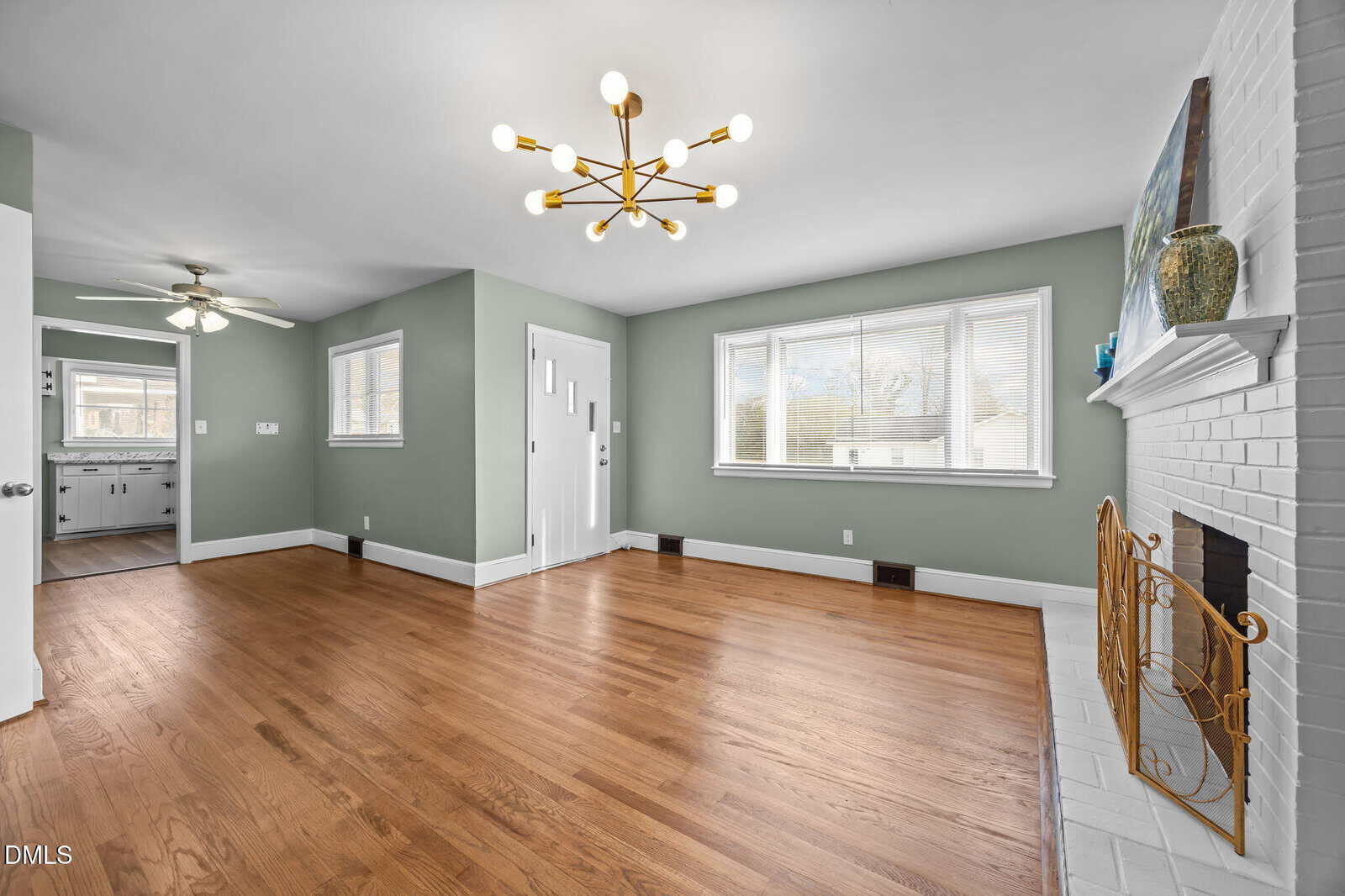 A bright living room with hardwood floors, sage green walls, a modern chandelier, large windows, and a white brick fireplace. A kitchen with white cabinetry is visible through an open doorway.
