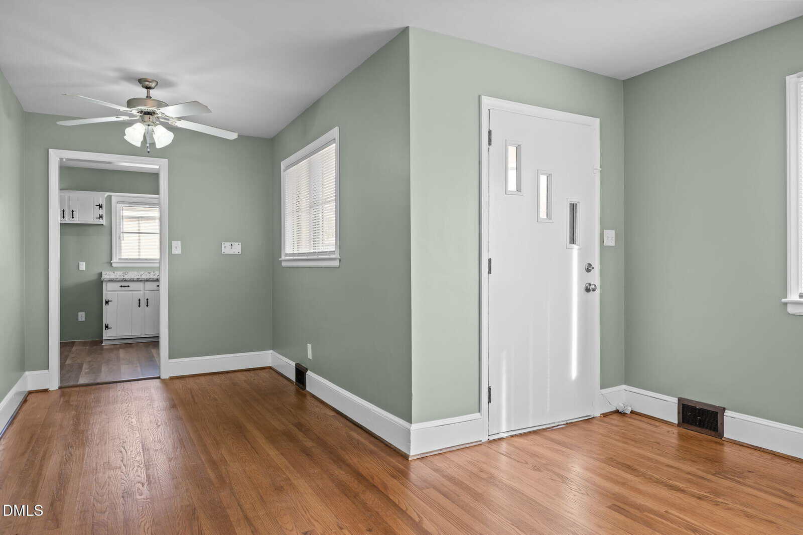 A small room with light green walls, hardwood floors, white trim, a white front door with three windows, ceiling fan, and view into a kitchen with white cabinets in the background.