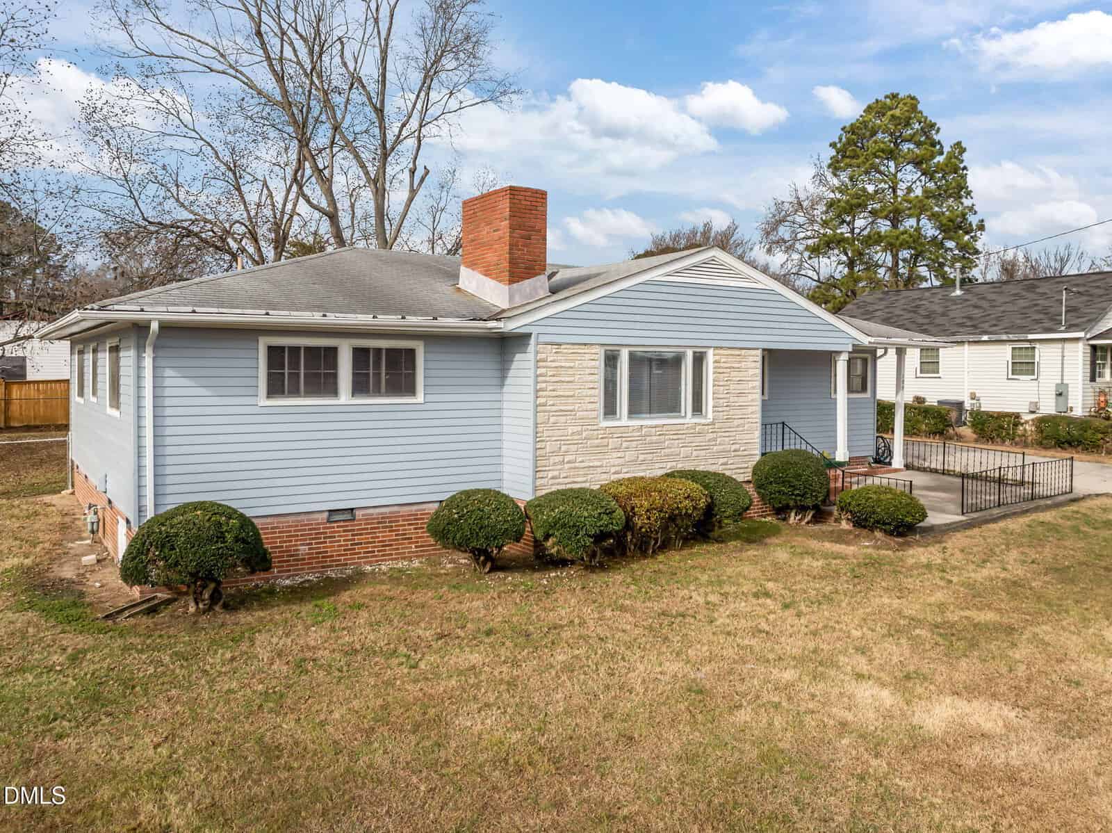 Single-story house with light blue siding, a brick chimney, and a stone accent wall in front. There are trimmed bushes along the foundation and a concrete walkway leading to the front steps. The yard is mostly grass.