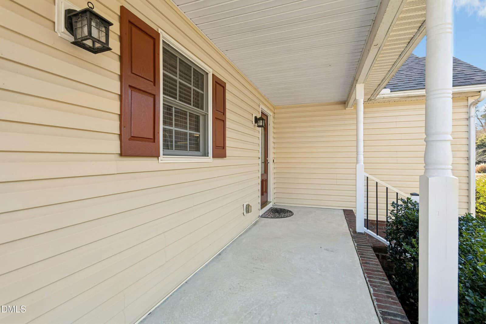 A covered front porch with cream-colored siding, a white ceiling, red shutters on the window, a black exterior light, and a brick-edged concrete floor leading to a front door with glass panels.