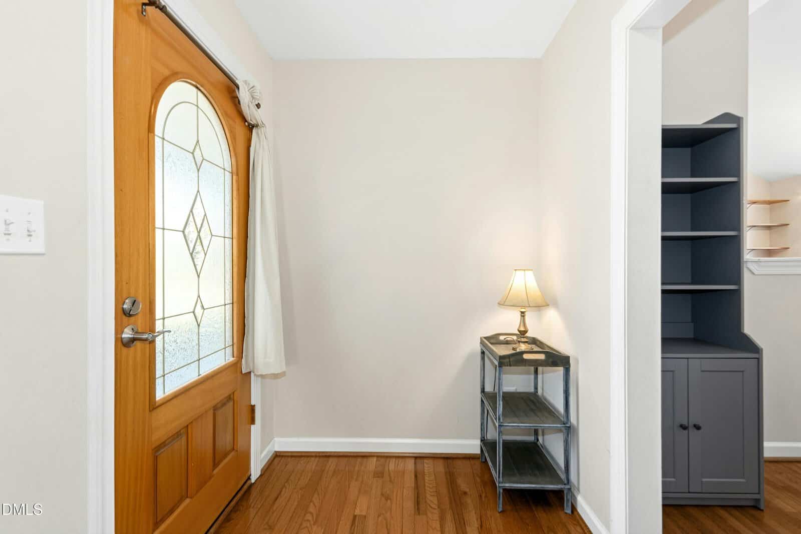 A small entryway with a wooden front door featuring a decorative glass panel, light curtains, hardwood floor, a gray side table with a lamp, and neutral-colored walls. Shelving is visible through an open doorway.