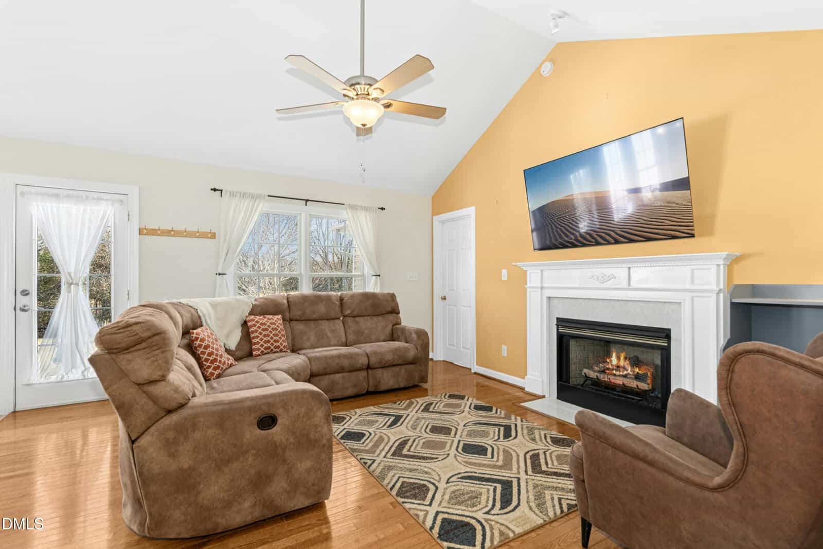 A bright living room with tan sectional sofas, a patterned rug, a fireplace with a TV above it, and a ceiling fan. Large windows and a glass door let in natural light, and the walls are painted white and yellow.