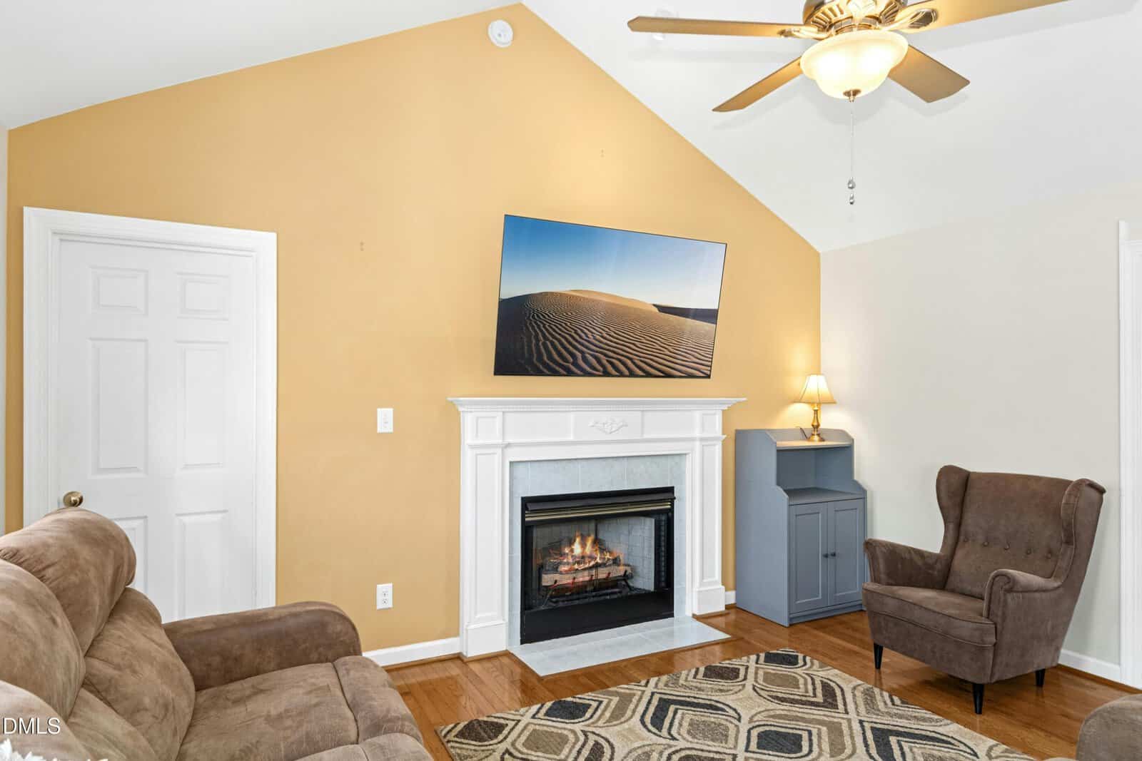 A cozy living room with a brown couch, an armchair, a fireplace with a TV mounted above it, a small cabinet with a lamp, and a ceiling fan. The walls are partly yellow with white trim and wood flooring.