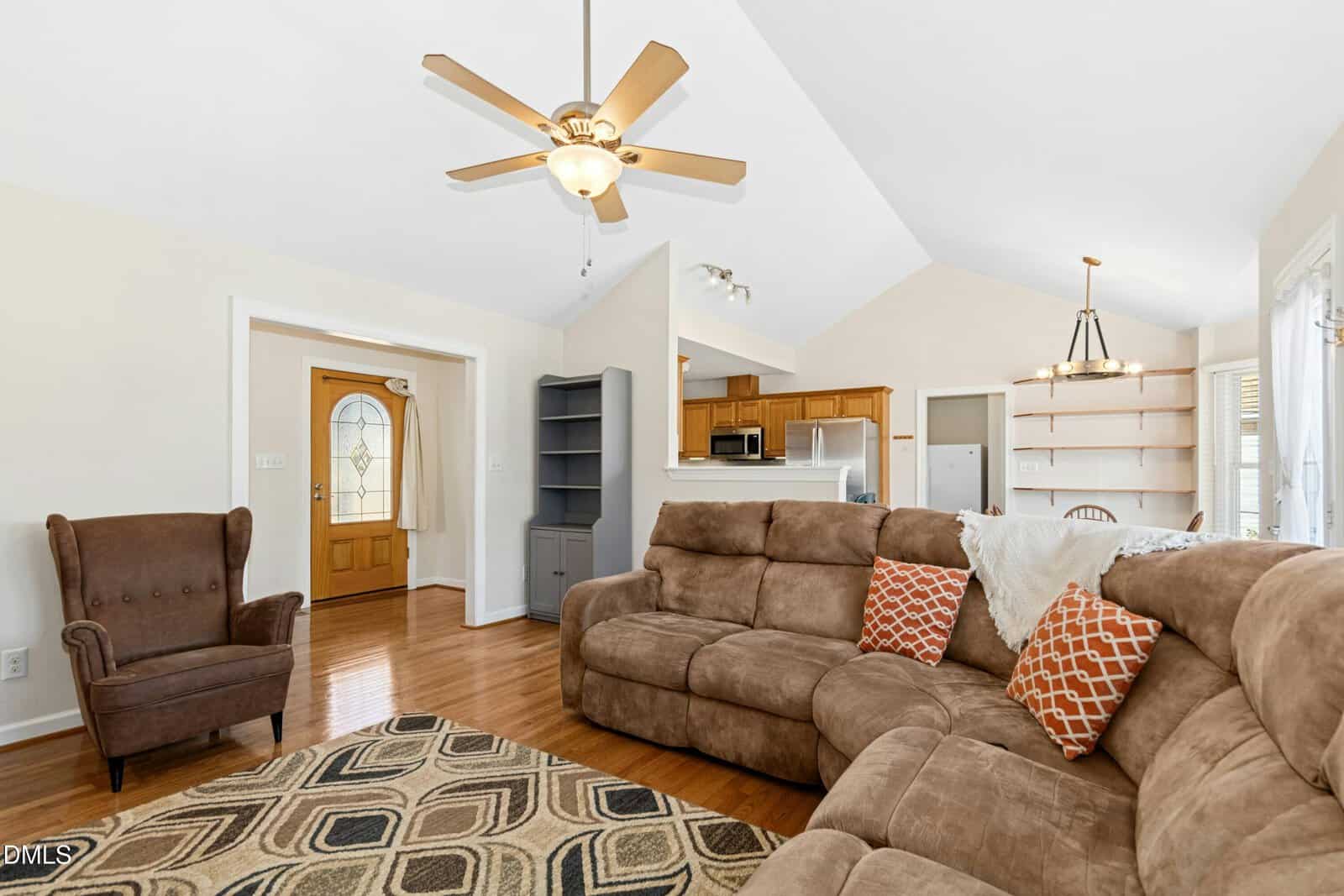 A cozy living room with brown sectional sofa, patterned rug, and armchair. Ceiling fan overhead, hardwood floors, and view into the kitchen with wooden cabinets and white appliances. Bright, airy space with neutral walls.