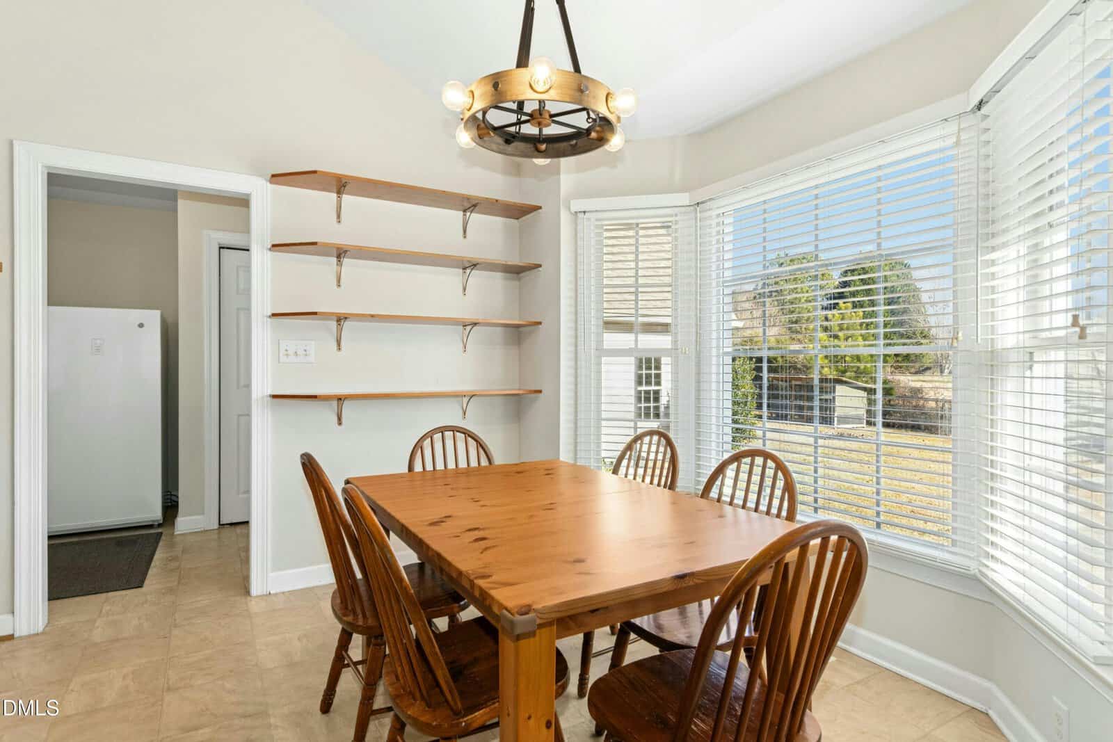 A bright dining area with a wooden table and four matching chairs, open wall shelves, a rustic chandelier, and large windows with white blinds overlooking a grassy backyard.