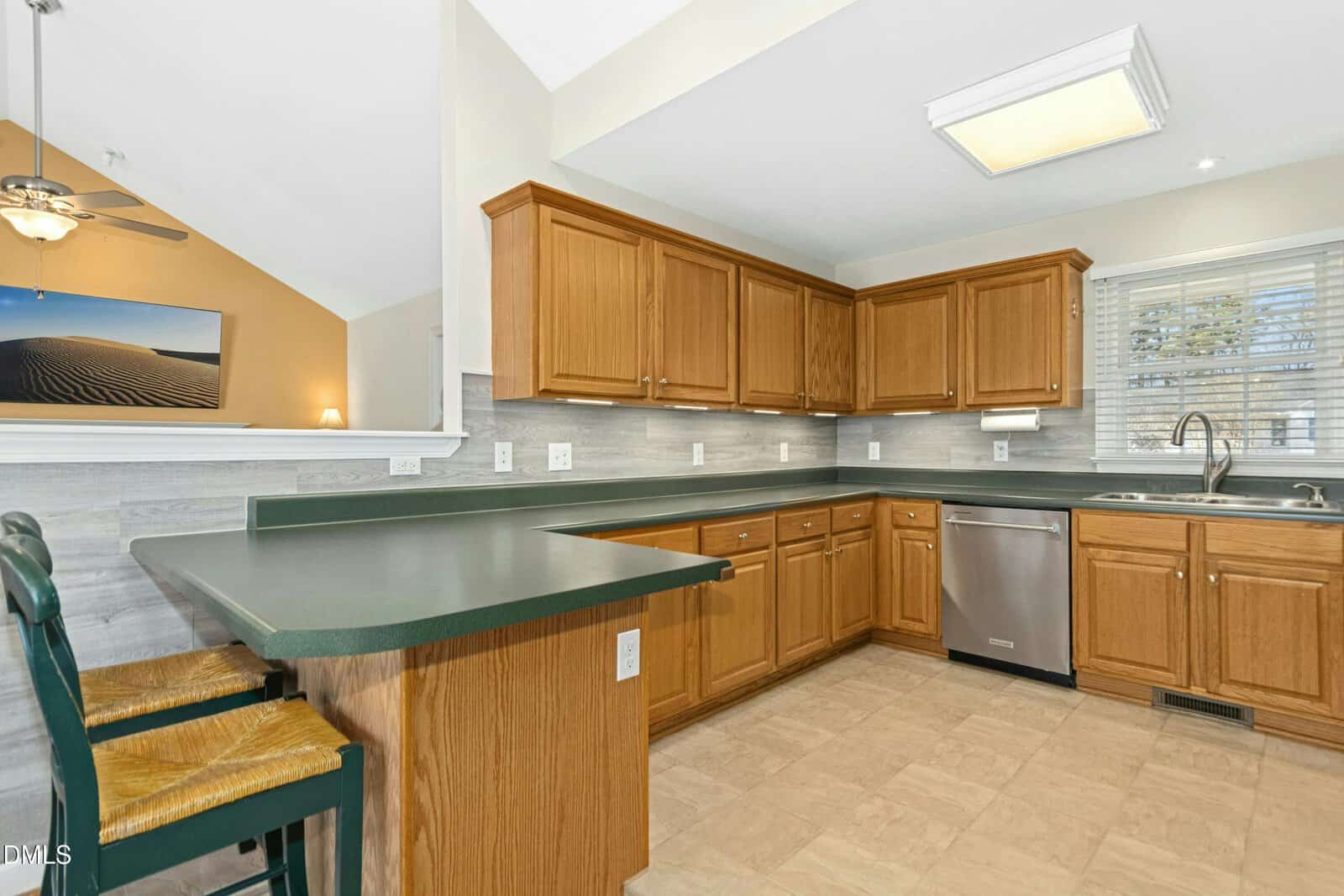 A spacious kitchen with wood cabinets, green countertops, stainless steel dishwasher, double sink under a window, bar seating with two chairs, and neutral tile flooring. There is an open view into the adjacent room.