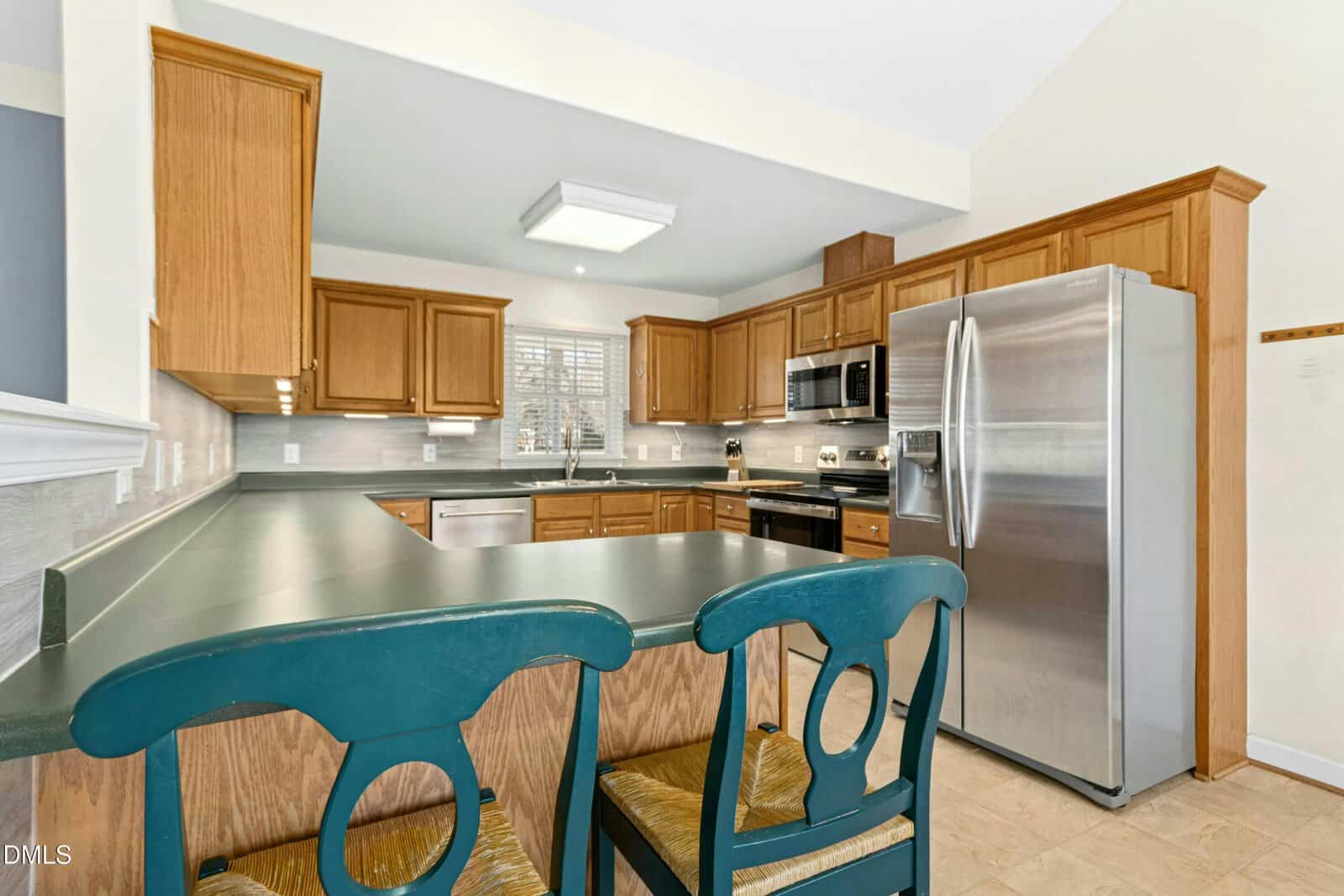 Kitchen with wood cabinets, green countertops, stainless steel appliances, and a large fridge. Two green chairs sit at the peninsula, and a window above the sink lets in natural light.