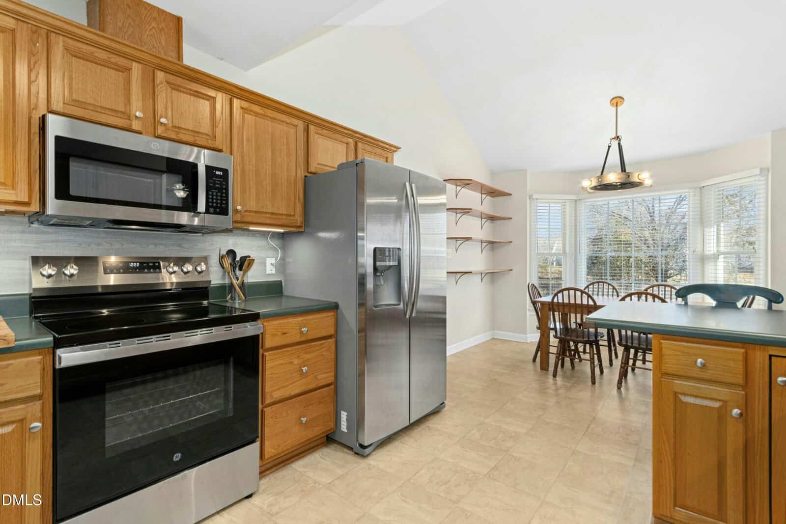 Kitchen with stainless steel appliances, wooden cabinets, green countertops, and a dining area with a wooden table and chairs by a bay window. Open shelves are mounted on the wall near the dining space.