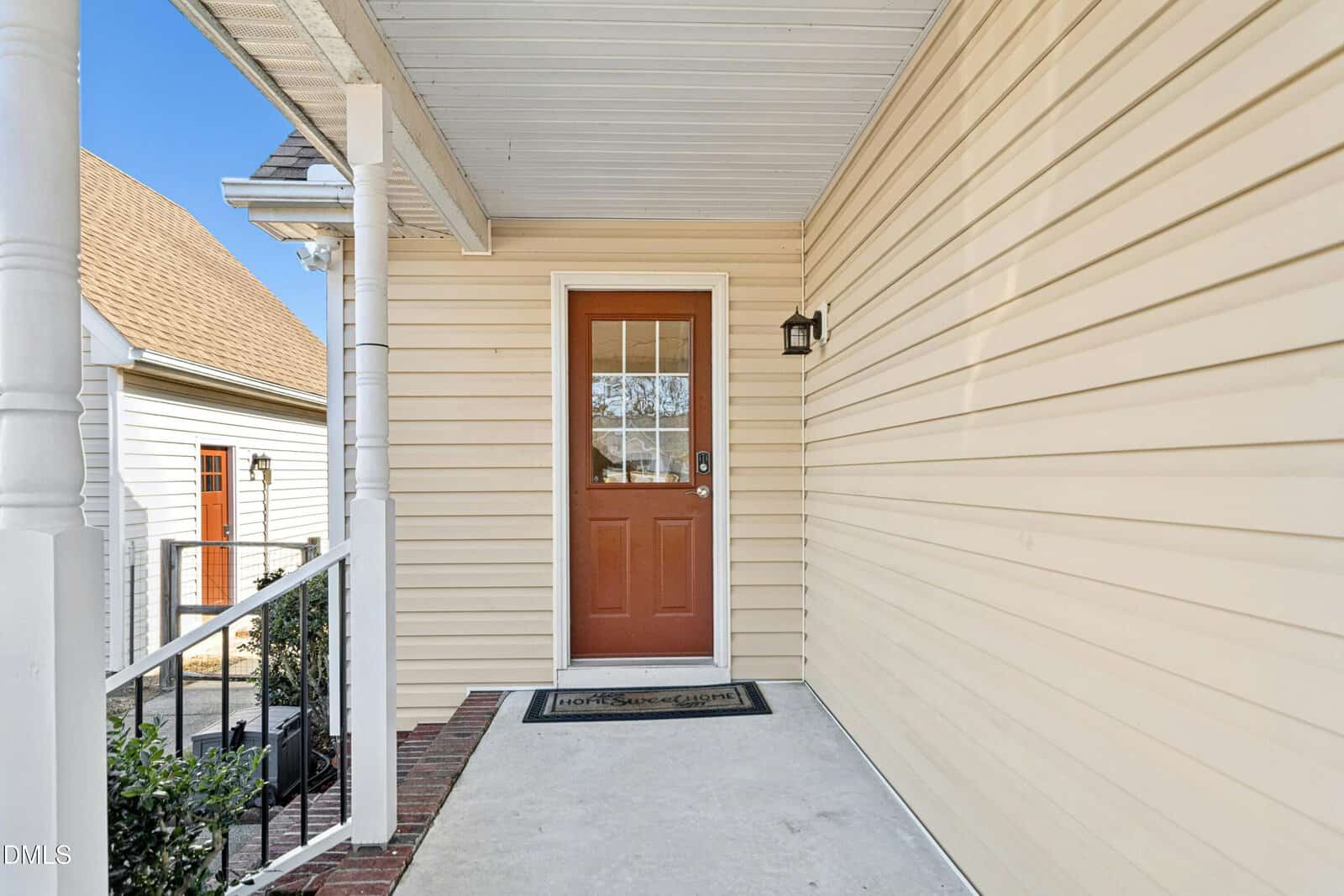 A covered front porch with beige vinyl siding, a brown door with glass window, a black wall-mounted light, a "Home Sweet Home" doormat, and black metal railing by the steps.