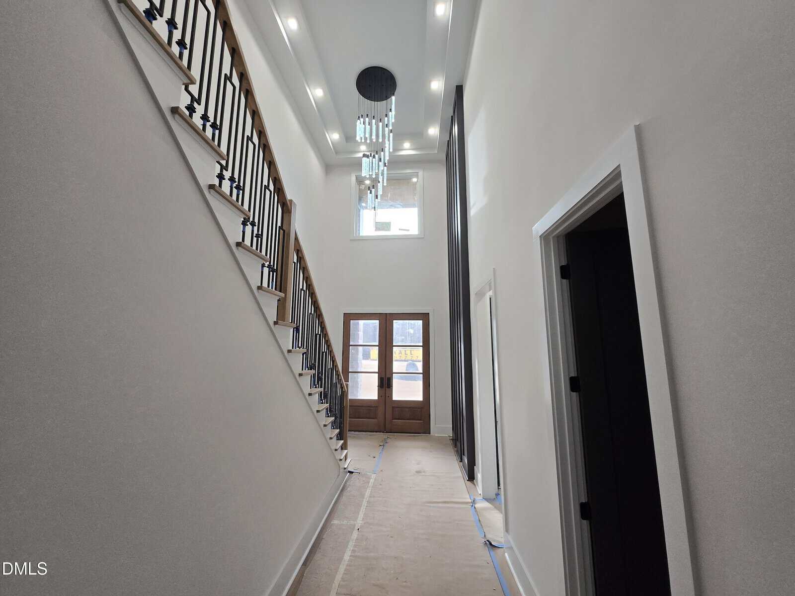 A modern, well-lit entry hallway with high ceilings, a large chandelier, wooden double front doors, and an open staircase with black railings on the left side. The floor is covered for protection.