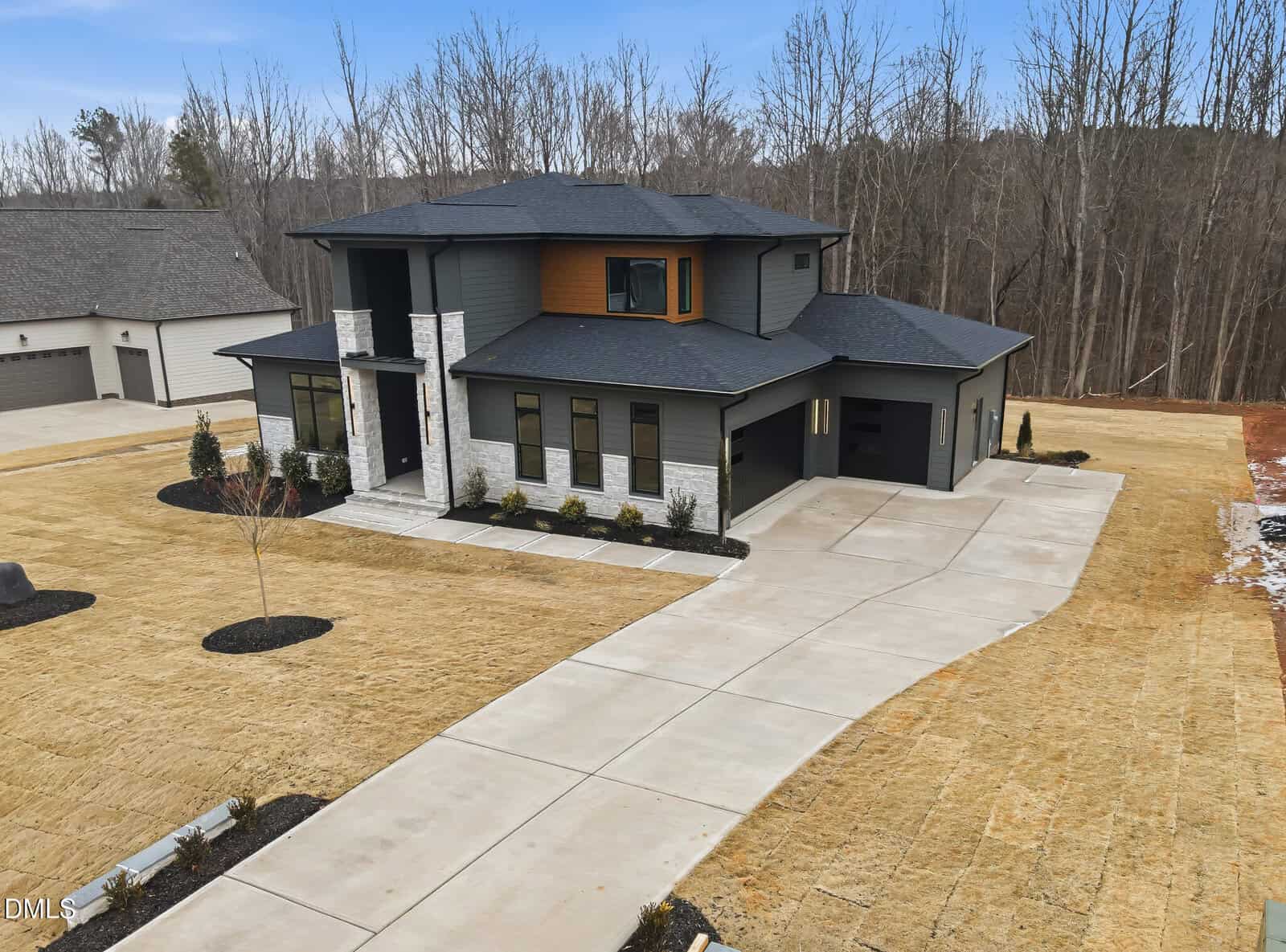 A modern two-story house with gray siding, large windows, and white stone accents, surrounded by sparse grass and a paved driveway, set next to a wooded area under a clear sky.
