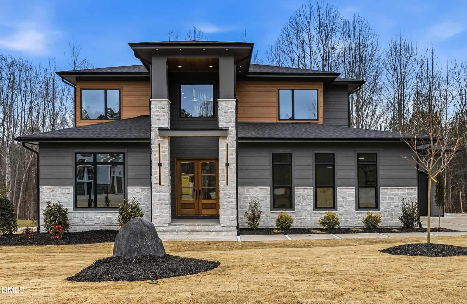 Modern two-story house with a mix of dark gray siding and stone exterior, large windows, a double front door, and a small porch, surrounded by a manicured lawn and a backdrop of leafless trees.