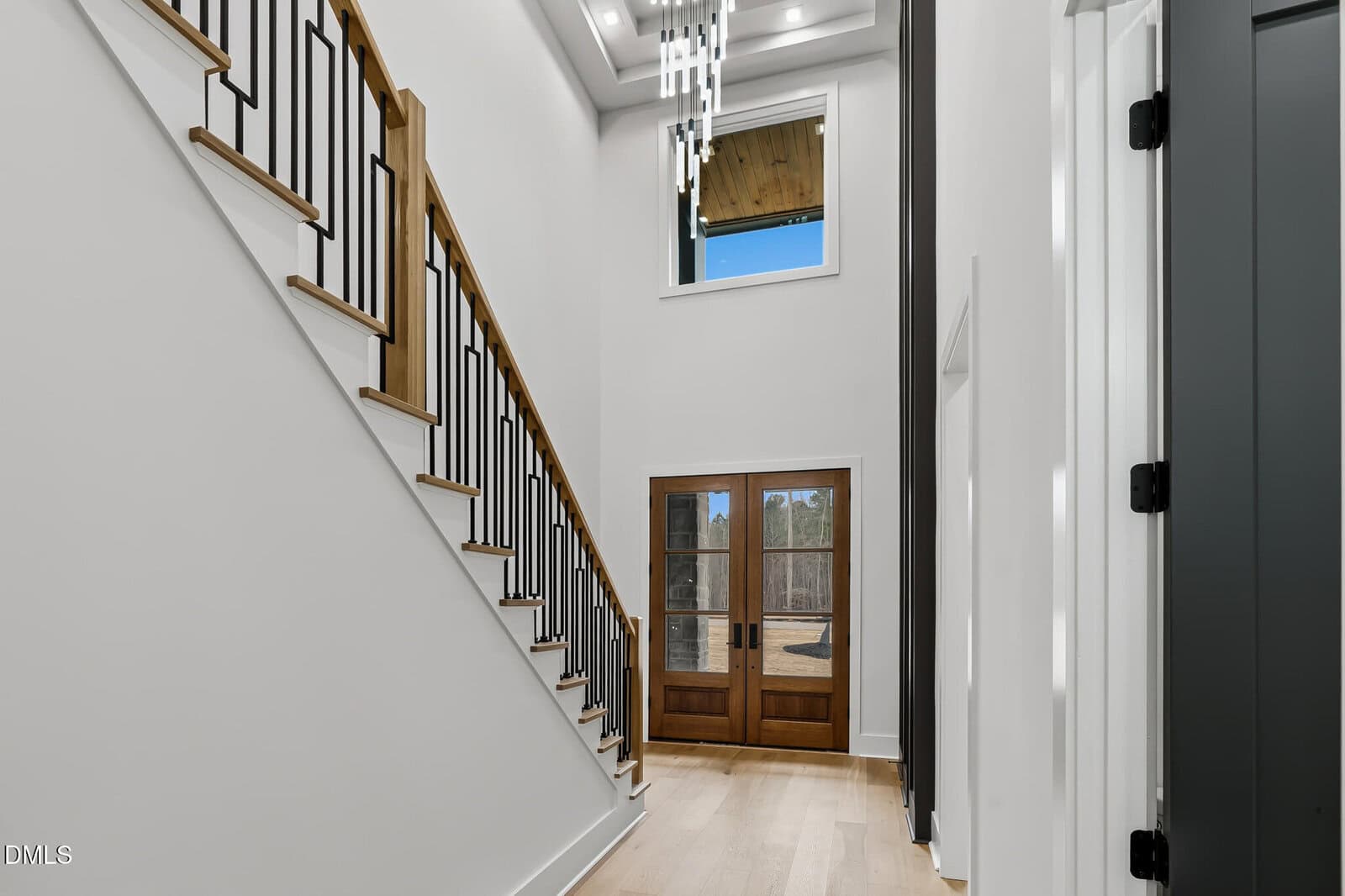 Modern entryway with light wood flooring, white walls, a wooden staircase with black railings on the left, and double wooden front doors with glass panes under a tall ceiling and contemporary chandelier.