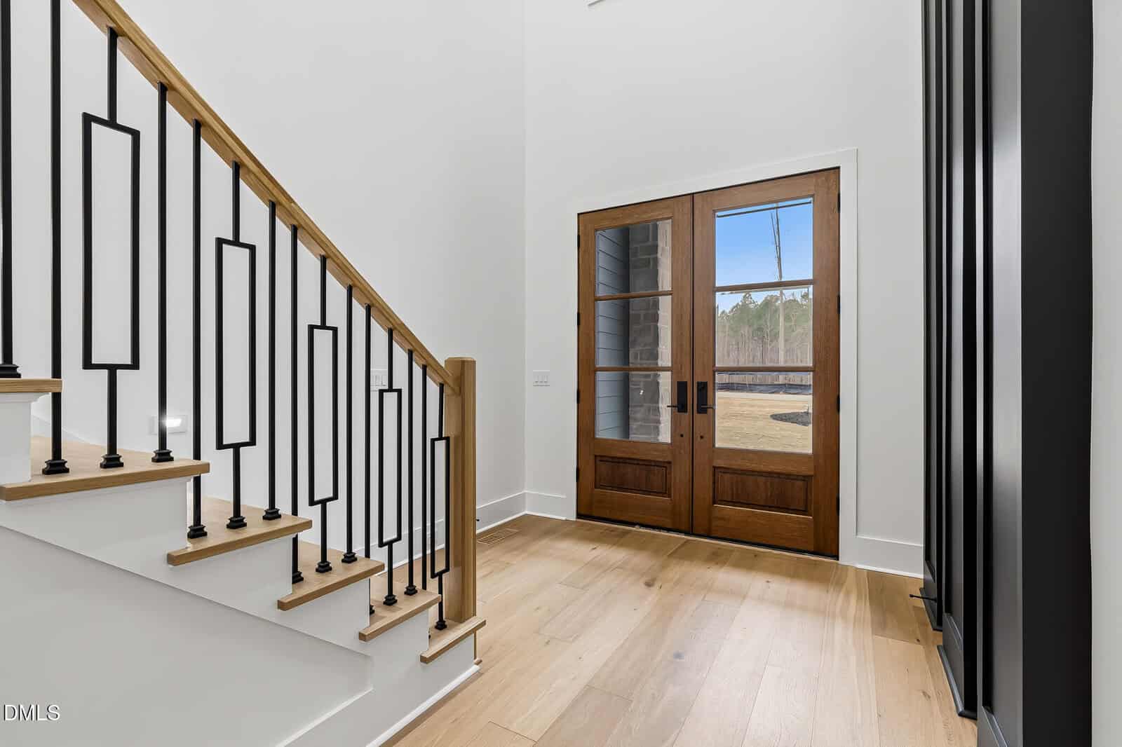 Bright entryway with light wood floors, white walls, a staircase with wooden handrail and black metal balusters, and double wooden front doors with large glass panels showing a view outside.