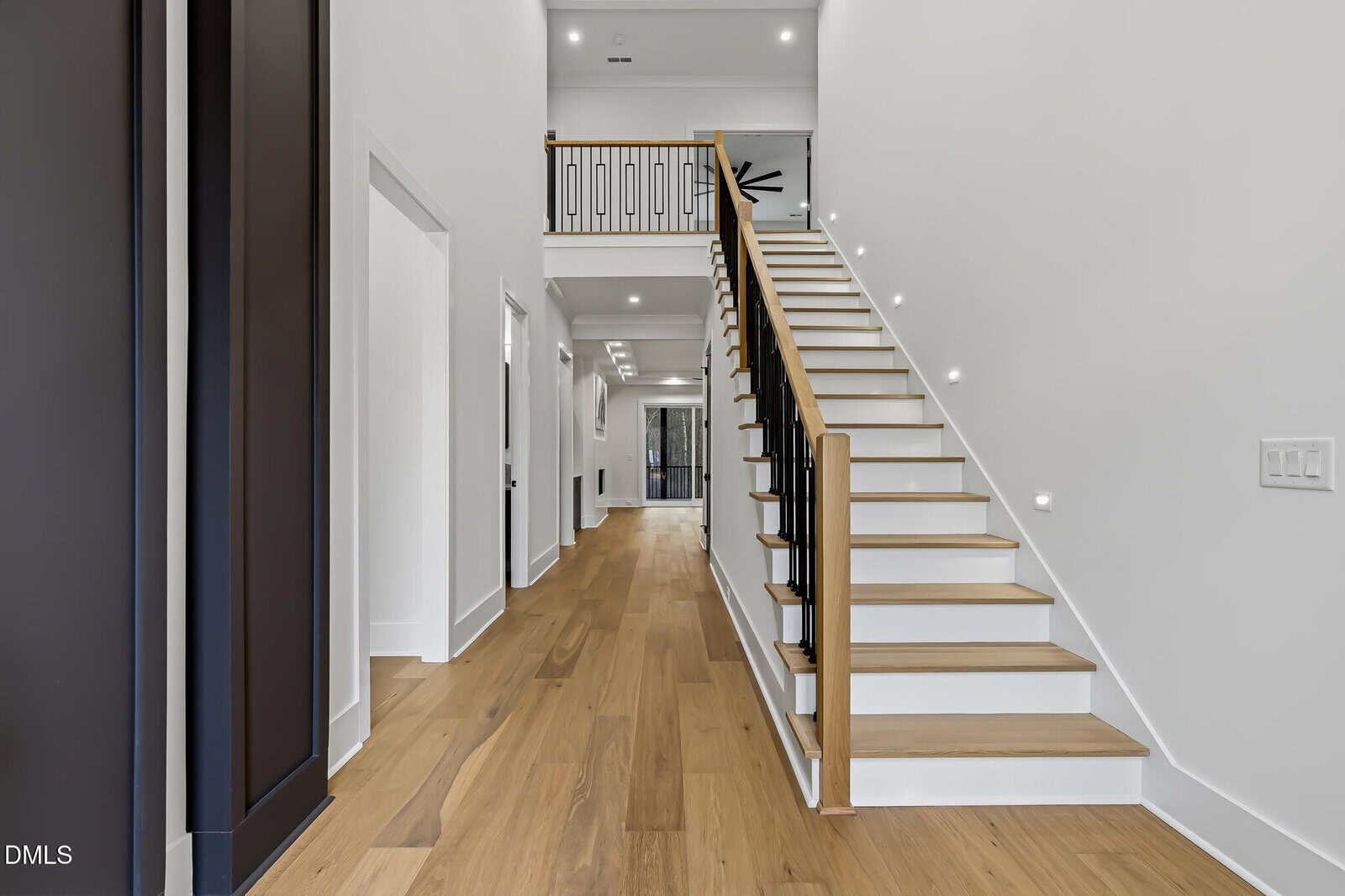 Modern home interior featuring a wooden staircase with black railings, white walls, and light wood flooring. A hallway extends to the left with doors on either side and a view of an upstairs balcony.