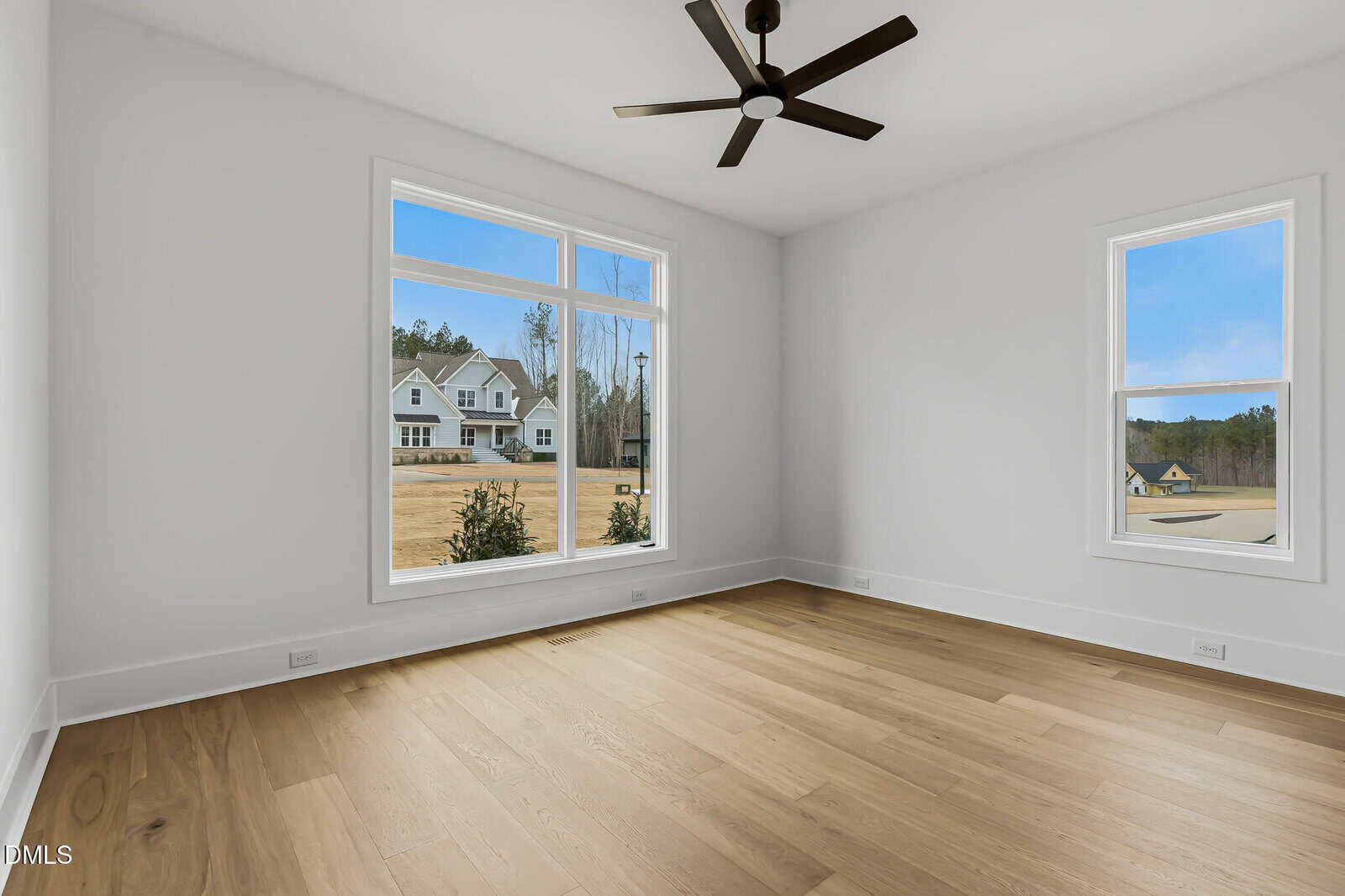Bright, empty room with light wood floors, white walls, two large windows showing houses and trees outside, and a black ceiling fan.