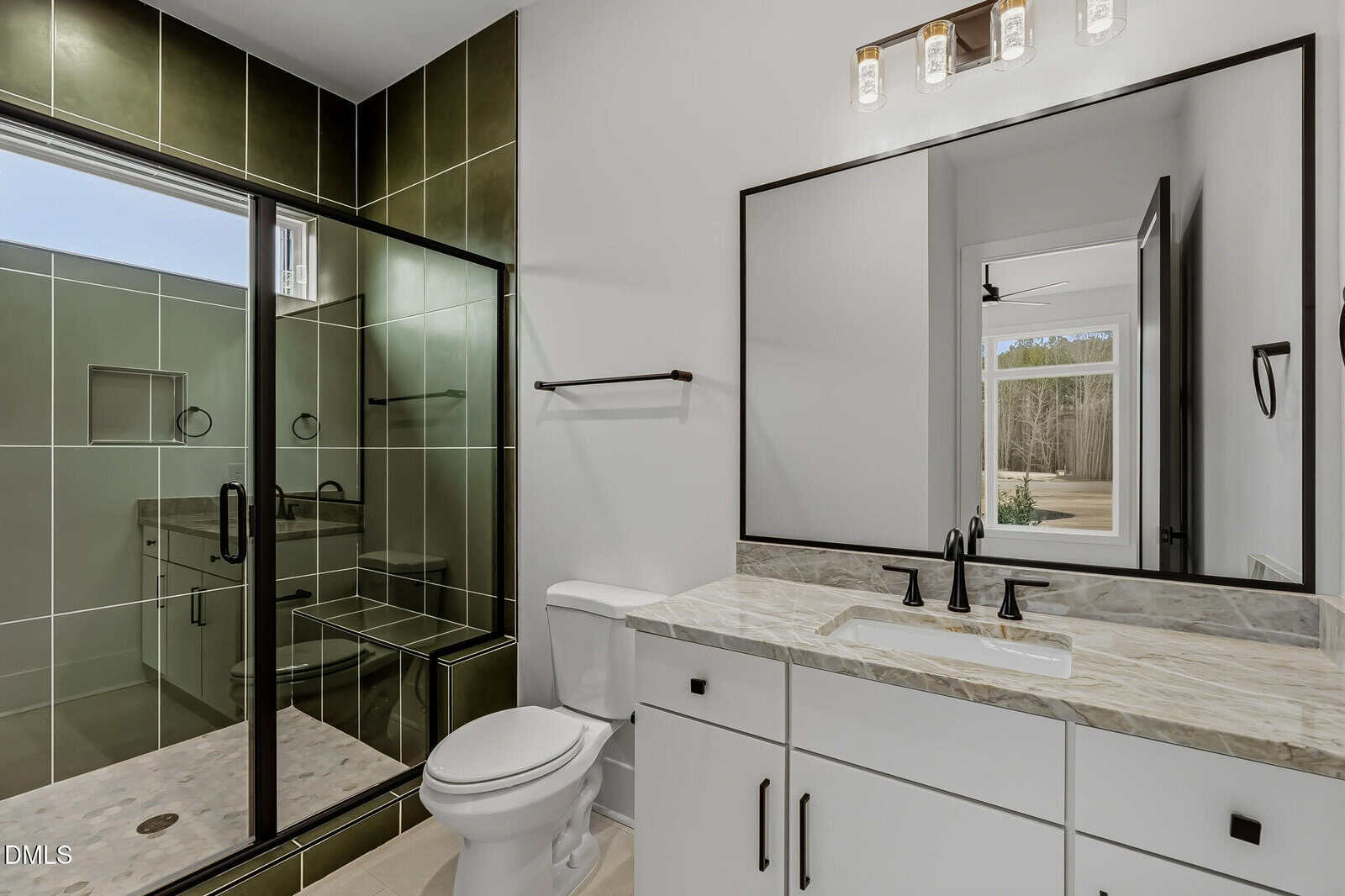Modern bathroom with a glass-enclosed shower featuring dark green tiles, a white toilet, a white vanity with a marble countertop, black fixtures, and a large mirror. A window brings natural light into the space.