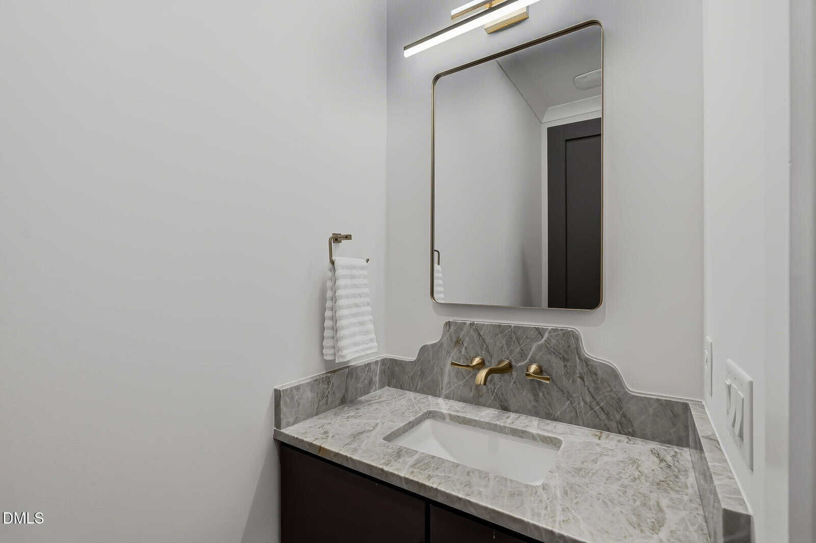 A modern bathroom vanity with a gray marble countertop, gold wall-mounted faucet, rectangular sink, large framed mirror, towel ring with a white towel, and white walls.