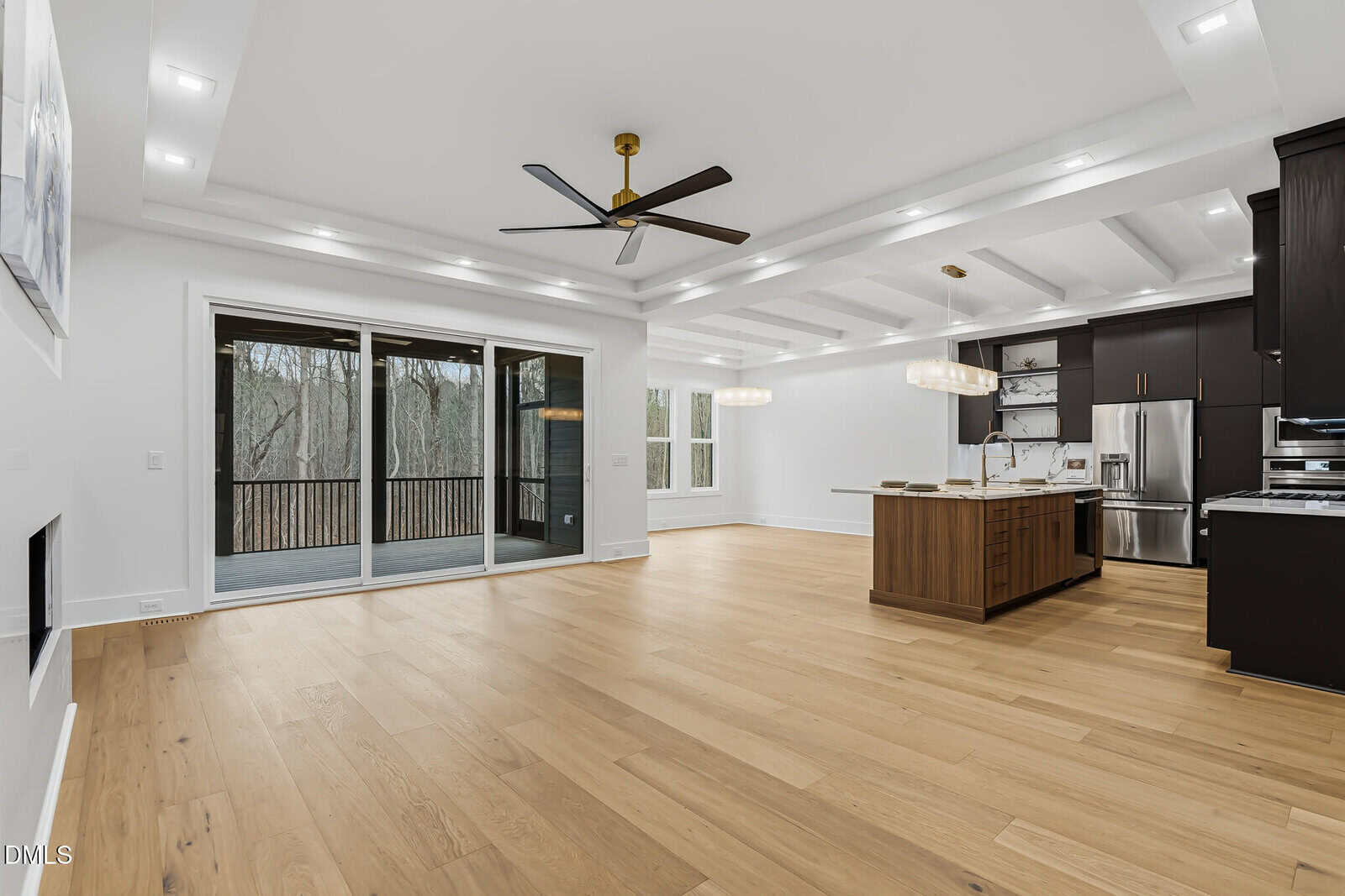 Spacious modern open-concept living area with light wood flooring, large sliding glass doors, a ceiling fan, recessed lighting, and a kitchen featuring a center island, stainless steel appliances, and dark cabinetry.