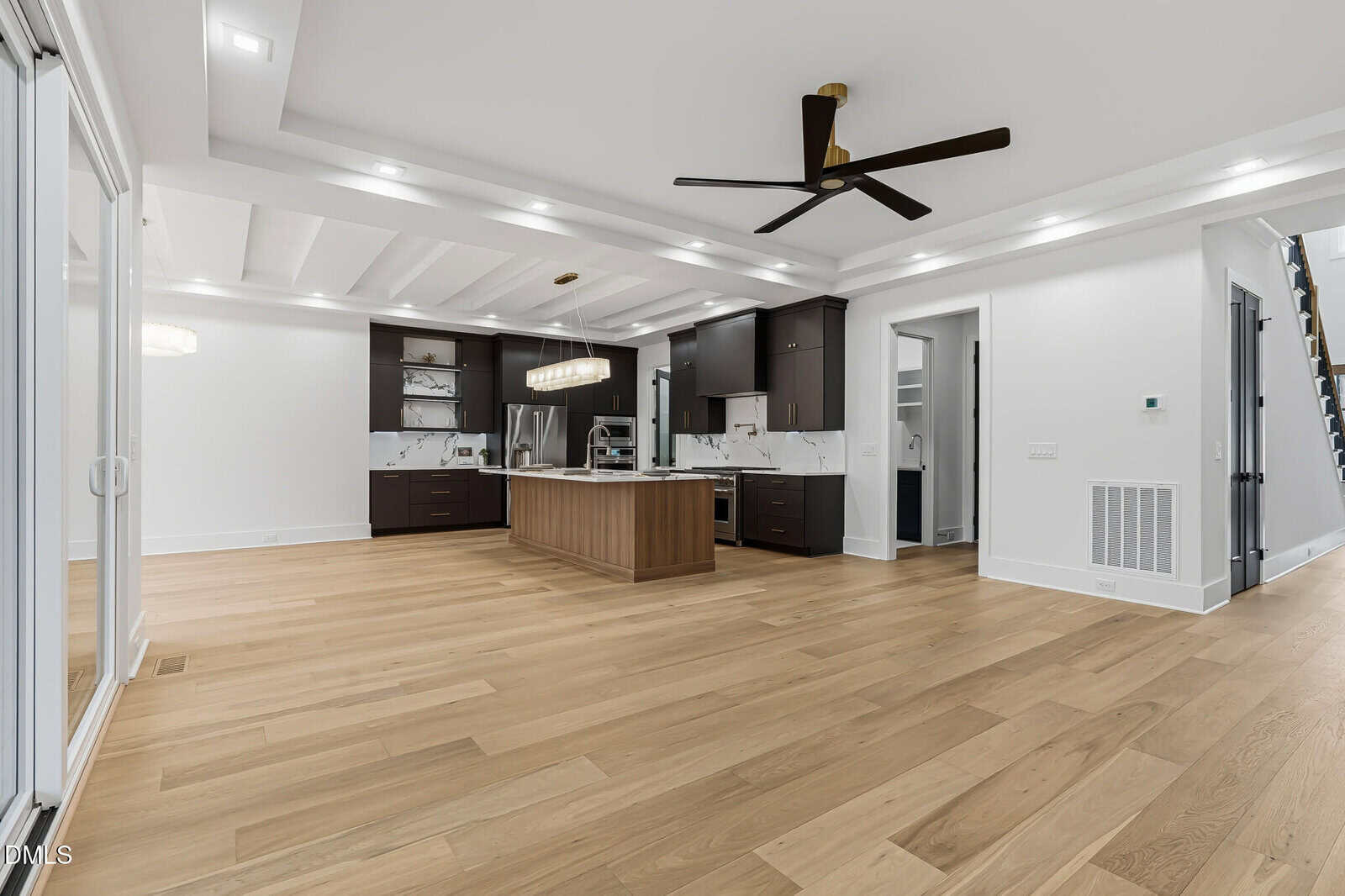 Spacious modern kitchen with light wood floors, dark cabinets, a center island, stainless steel appliances, and a ceiling fan. White walls and recessed lighting create a bright, open atmosphere.