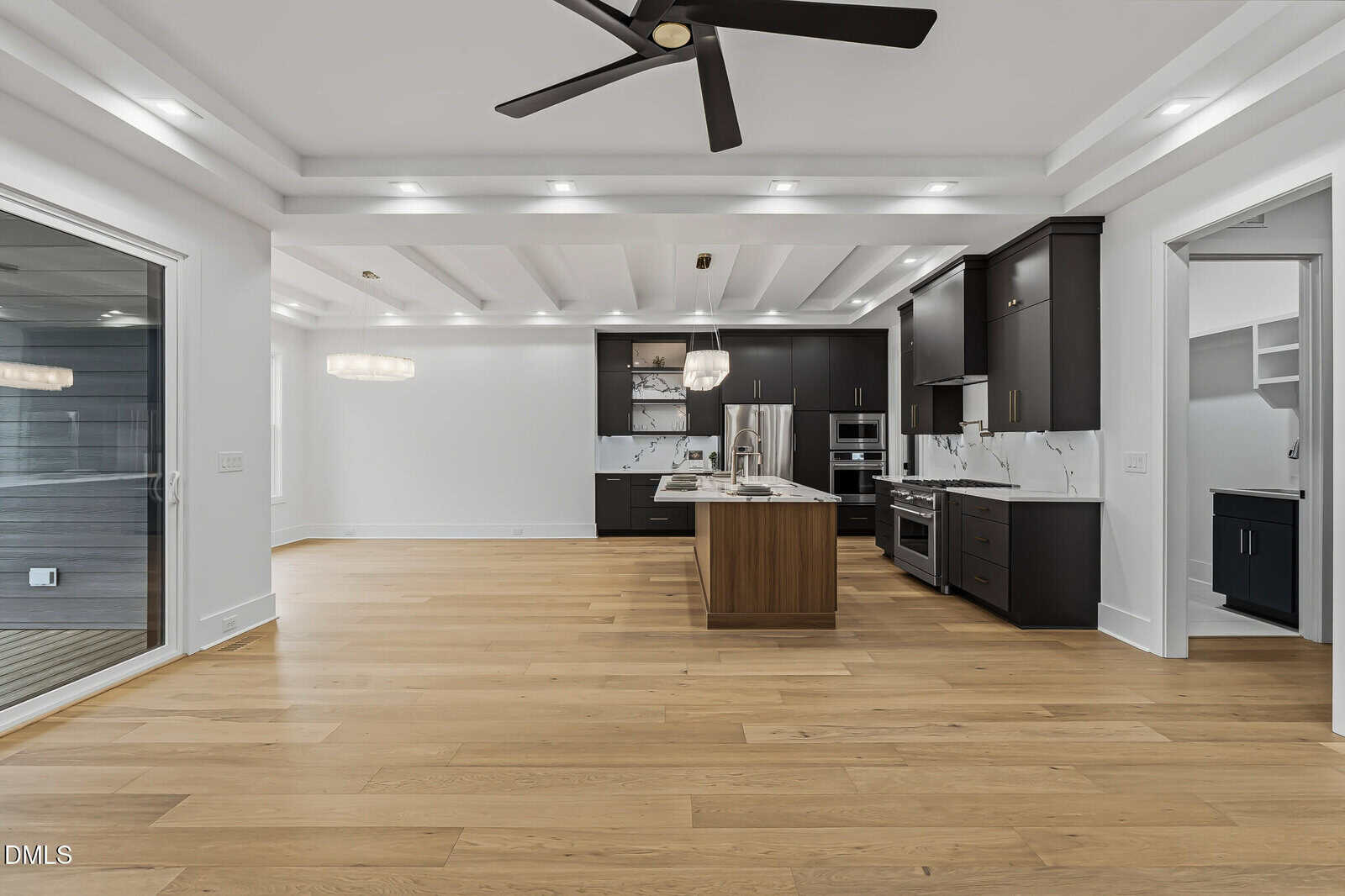 Modern kitchen with black cabinets, stainless steel appliances, an island with wood finish, light wood flooring, recessed lighting, and a ceiling fan. Adjacent rooms and sliding glass door are also visible.