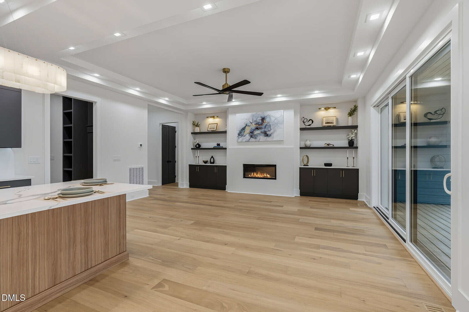 Modern living room with light wood floors, built-in shelves, a linear fireplace below abstract art, black cabinetry, recessed lighting, a ceiling fan, and sliding glass doors leading to an outdoor space. Kitchen island in foreground.
