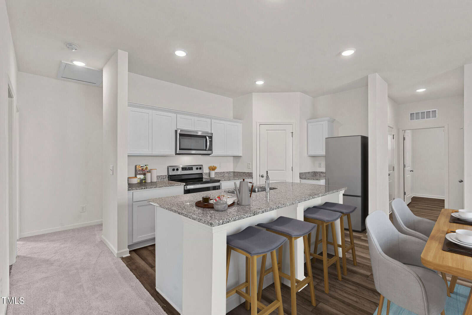 A modern kitchen with white cabinets, stainless steel appliances, and granite countertops. There are three gray barstools at the island and a dining table with four chairs on the right. The flooring is a mix of carpet and wood.