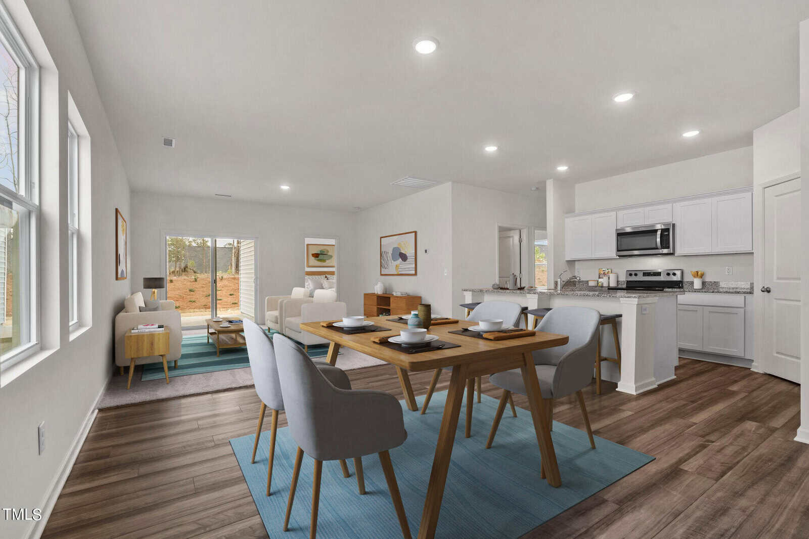 A modern open-concept living and dining area with white walls, wood flooring, light gray and wood furniture, and a kitchen featuring white cabinets and stainless steel appliances. Large windows let in natural light.