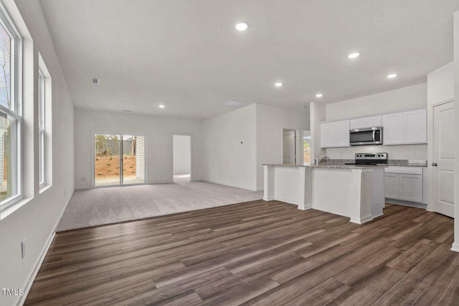 Open-plan living area with hardwood floors, adjoining carpeted space, and a kitchen featuring white cabinets, granite countertops, and stainless-steel appliances; large windows and sliding glass doors provide natural light.