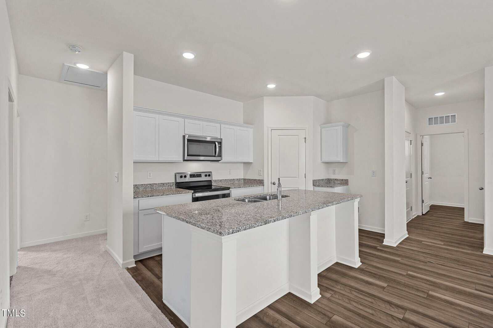 A modern kitchen with white cabinets, stainless steel appliances, granite countertops, a center island with a sink, and wood-style flooring. The room is brightly lit with recessed ceiling lights.