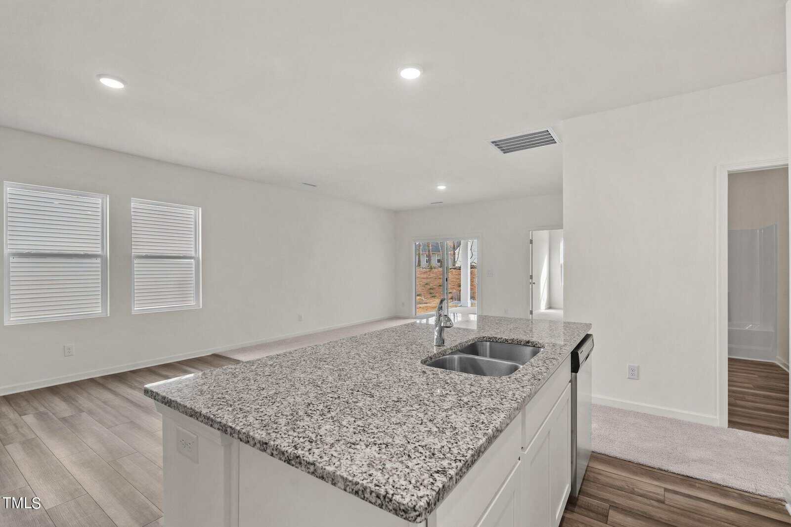 A modern, open-concept living space with a granite kitchen island in the foreground, wood-style flooring, white walls, and large windows. A sliding glass door and a doorway to a bathroom are visible in the background.