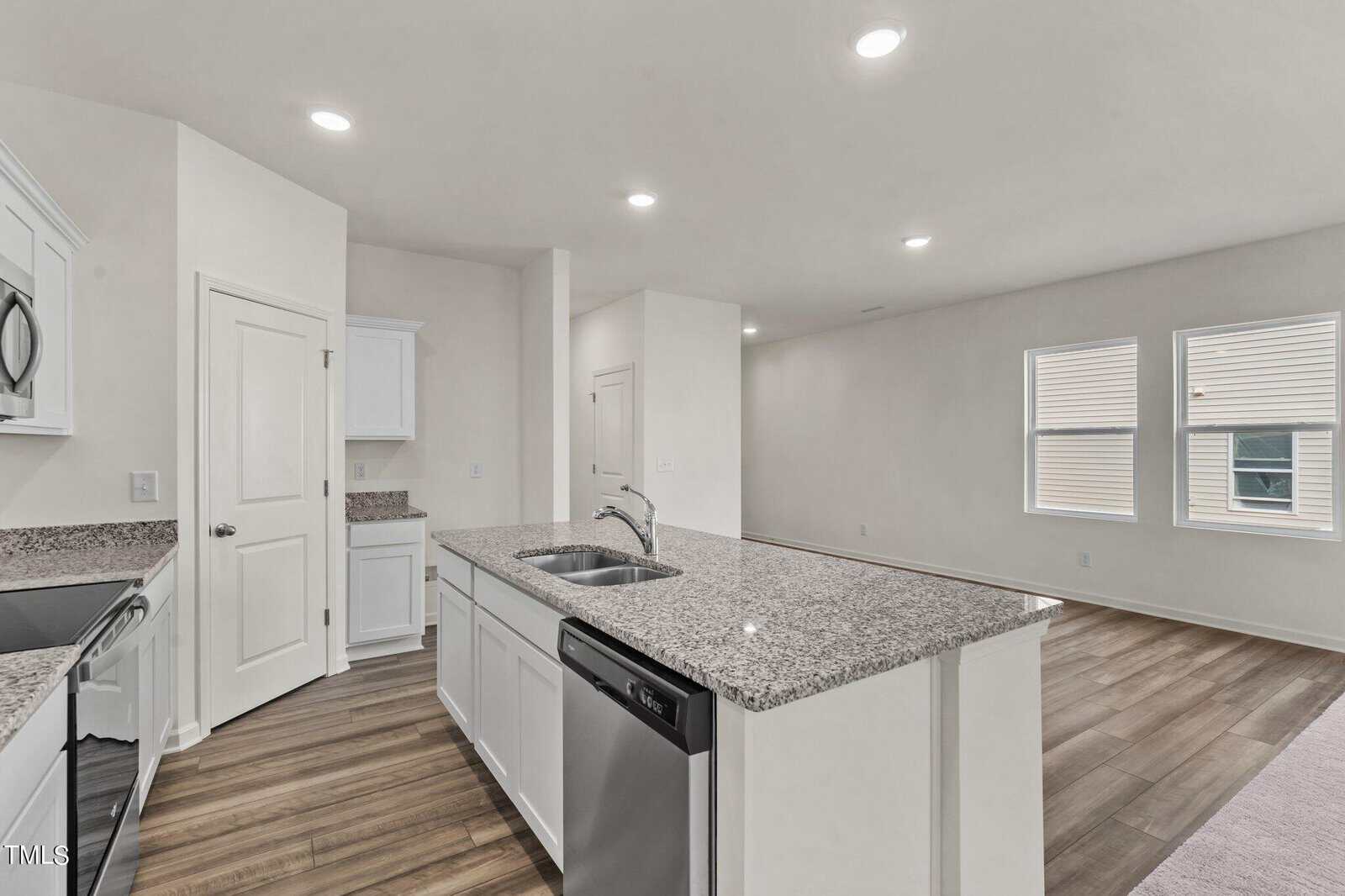 A modern kitchen with white cabinets, granite countertops, stainless steel appliances, and a double sink, open to a living area with wood flooring and large windows. Walls are painted light beige.