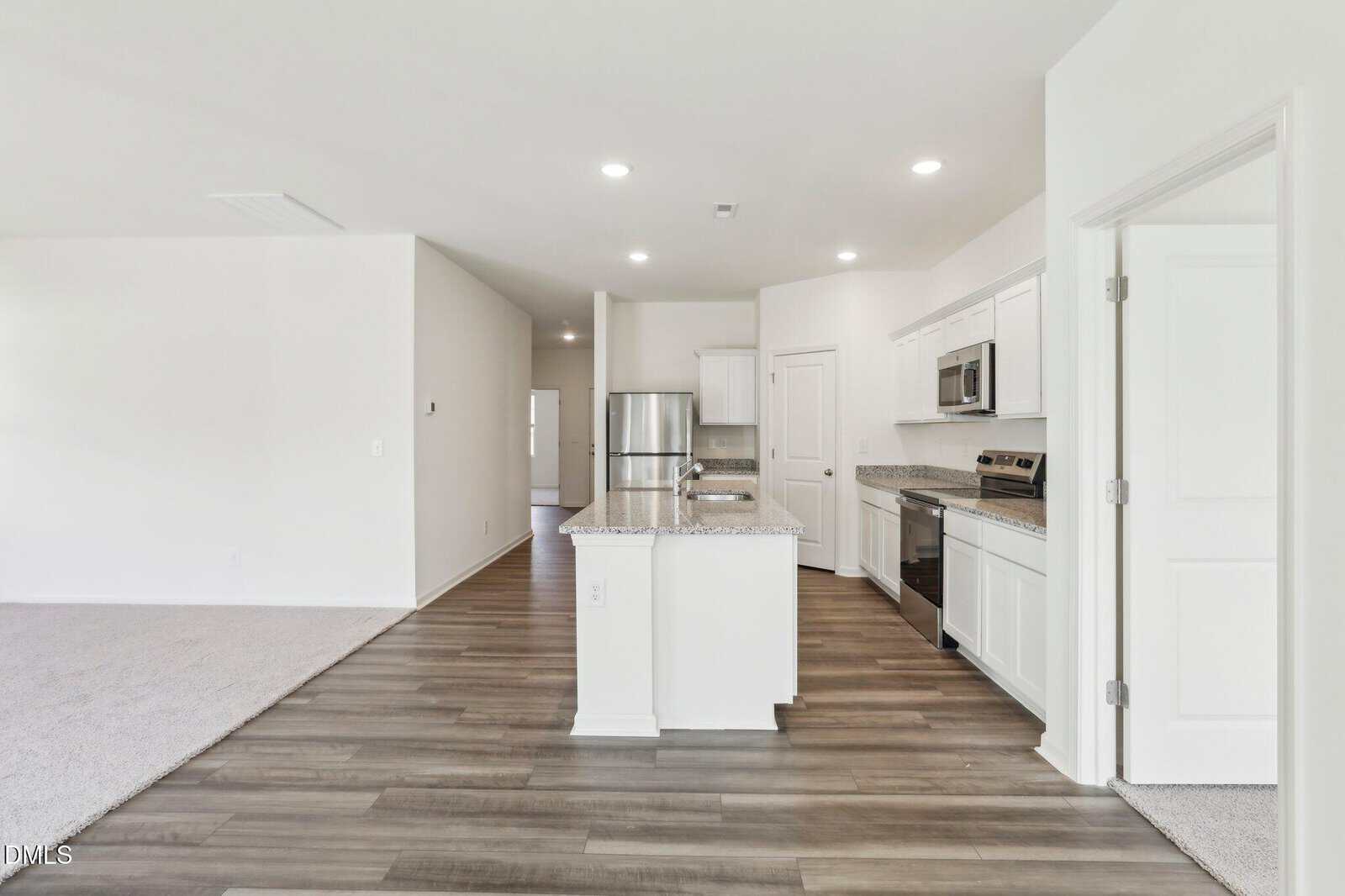 Modern open-concept kitchen with white cabinets, stainless steel appliances, and an island. Adjacent to a carpeted living area and hallway, with wood-style flooring in the kitchen. Walls are painted white.