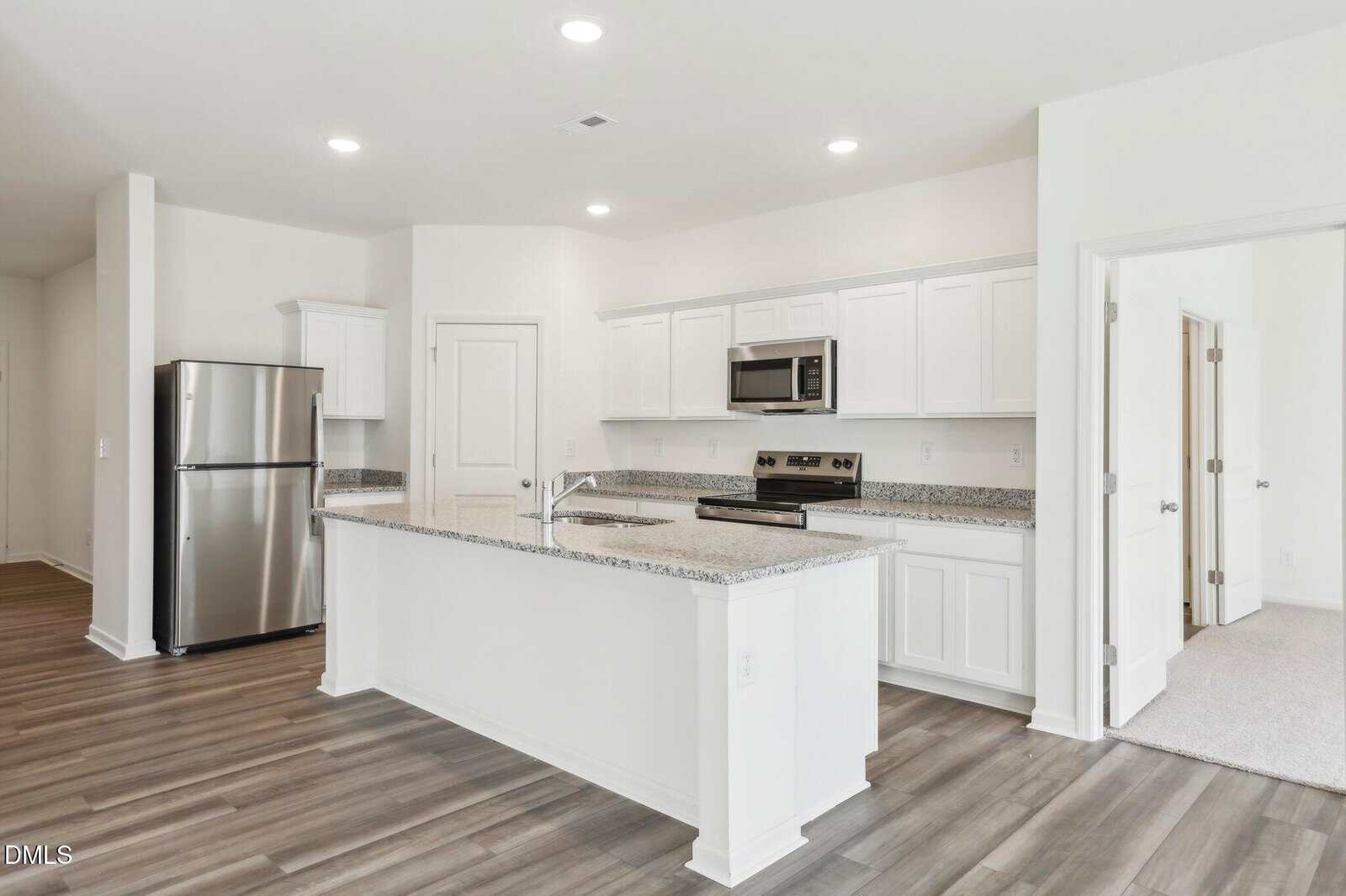 Modern kitchen with white cabinets, stainless steel appliances, granite countertops, large island, and wood-look flooring. Two doors lead to adjacent rooms; the space is bright with recessed lighting and a neutral color palette.