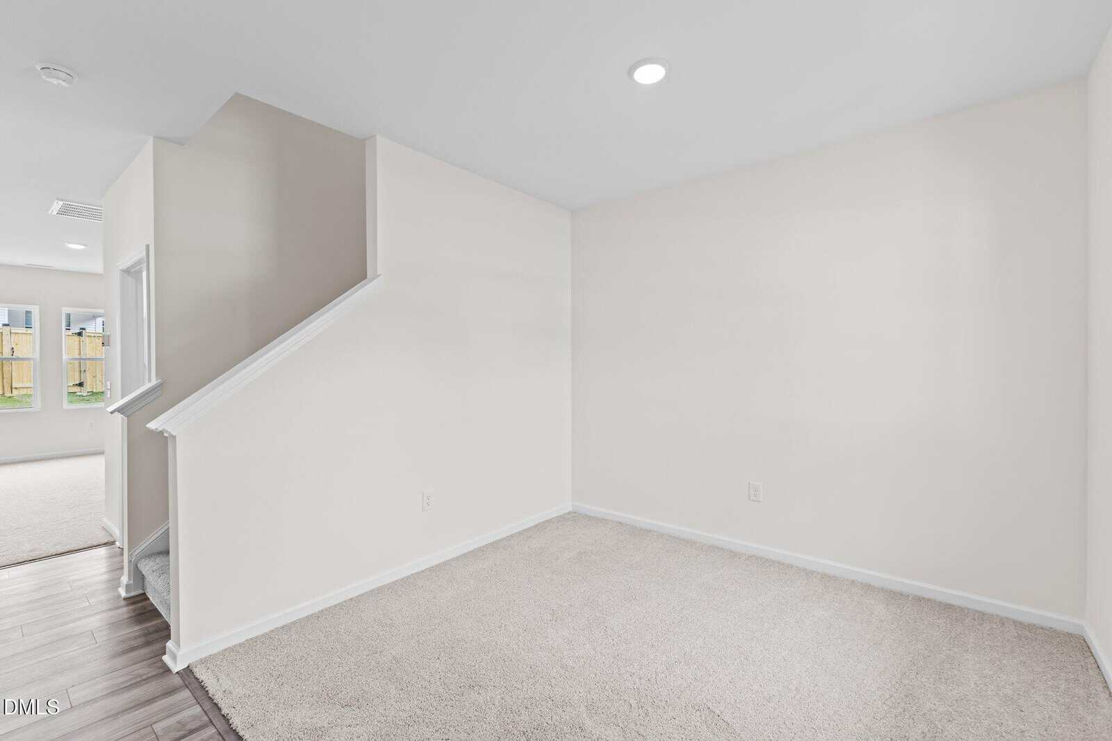 Empty room with beige carpet, white walls, and a partial view of wooden stairs with a white railing. Recessed ceiling light is on, and a doorless entry leads to another room with a window and wood flooring.