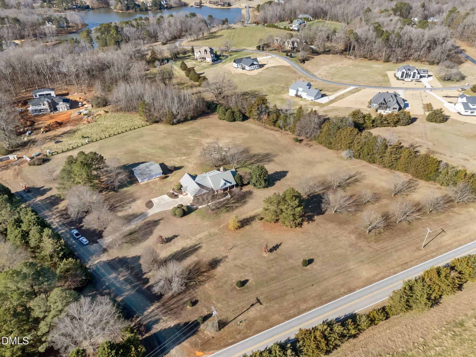 Aerial view of a large house with a circular driveway, surrounded by trees and open grassy land, set in a rural neighborhood with several other homes and a lake in the background.