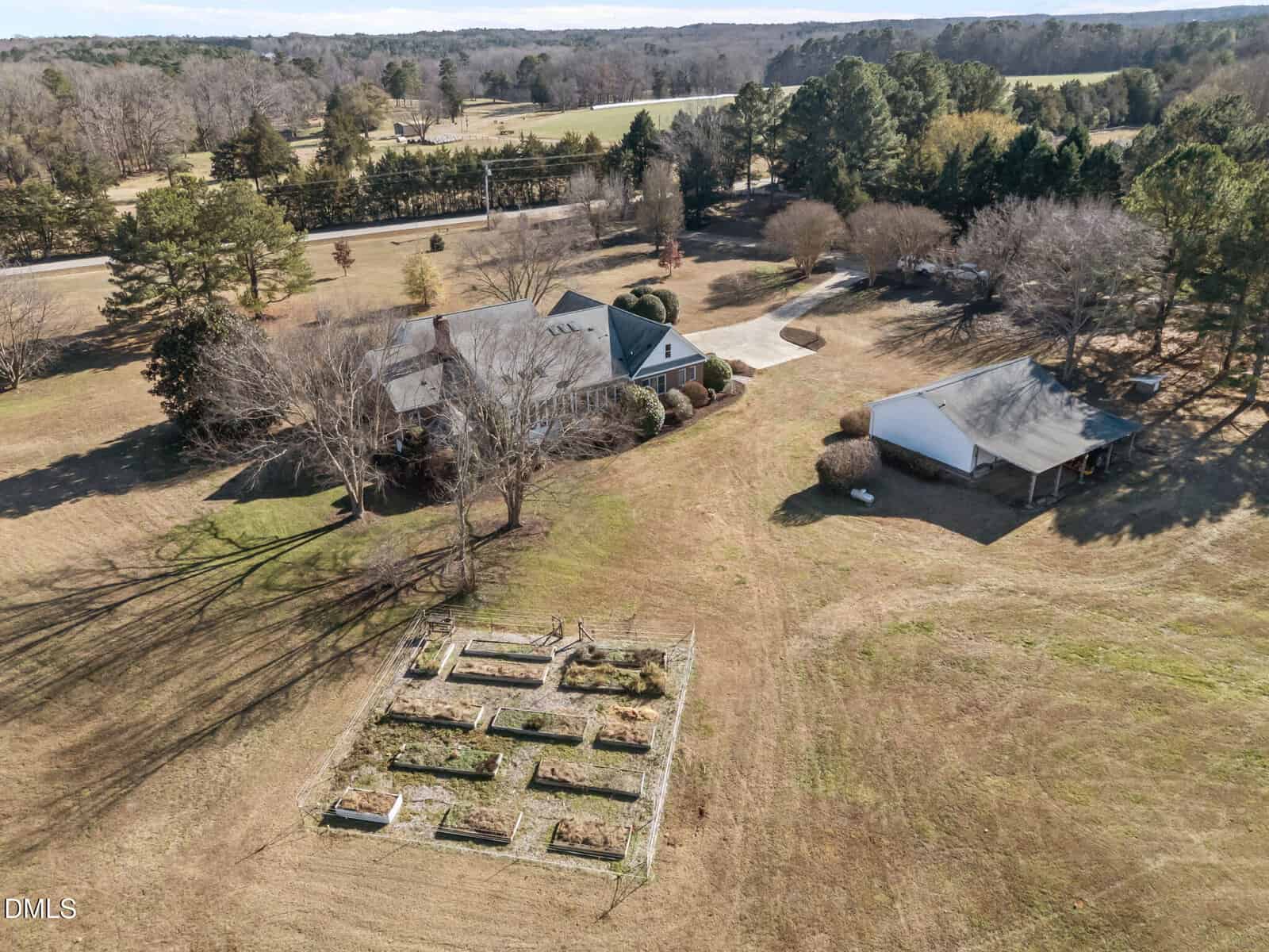 Aerial view of a house with a green roof, a detached garage, and a fenced vegetable garden with raised beds on a large grassy property surrounded by trees.