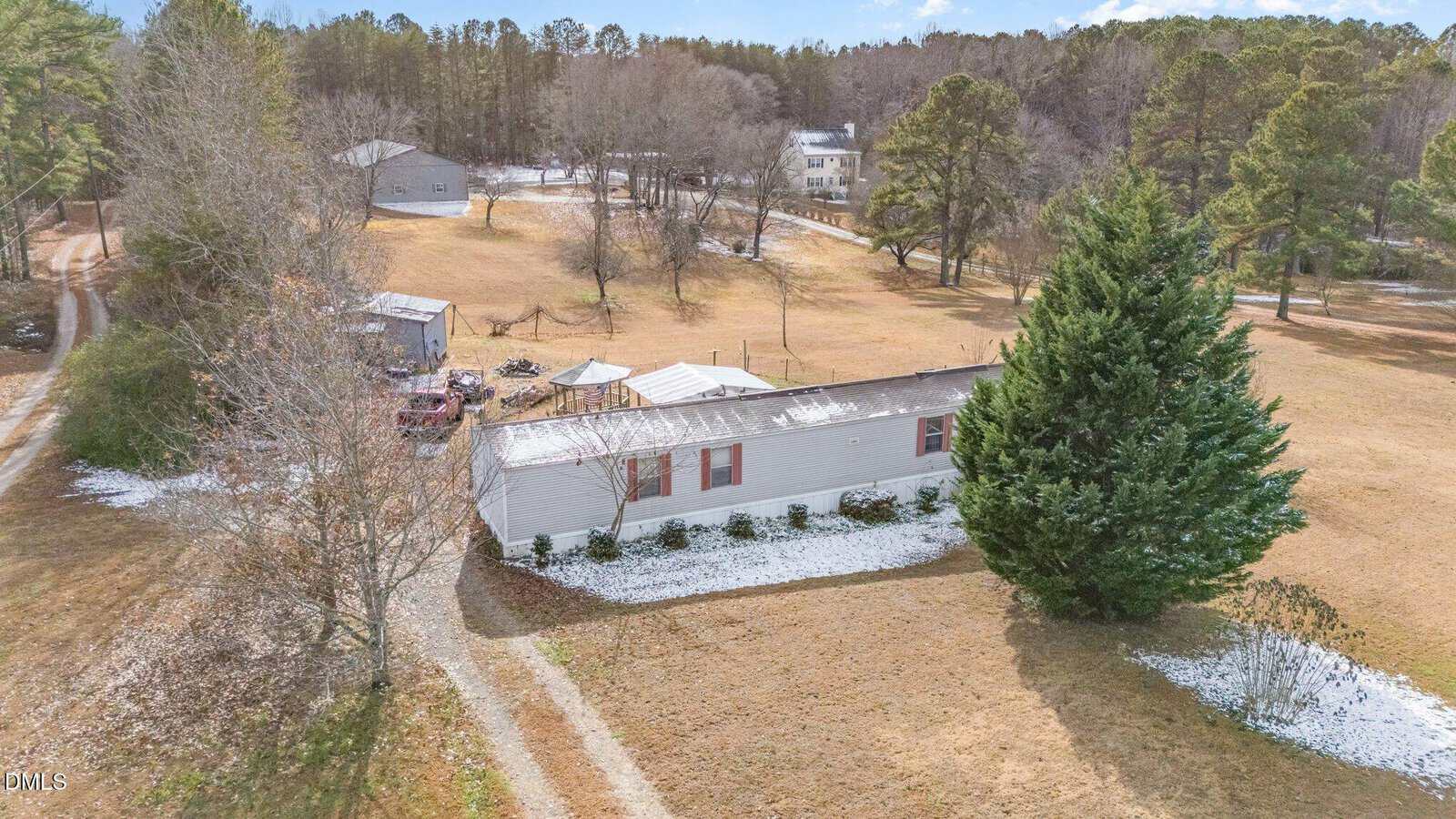 A single-story mobile home with a light-colored exterior sits on a spacious plot with patchy snow on the ground. There are trees, several outbuildings, and a gravel driveway leading to the home. Other houses are visible in the background.