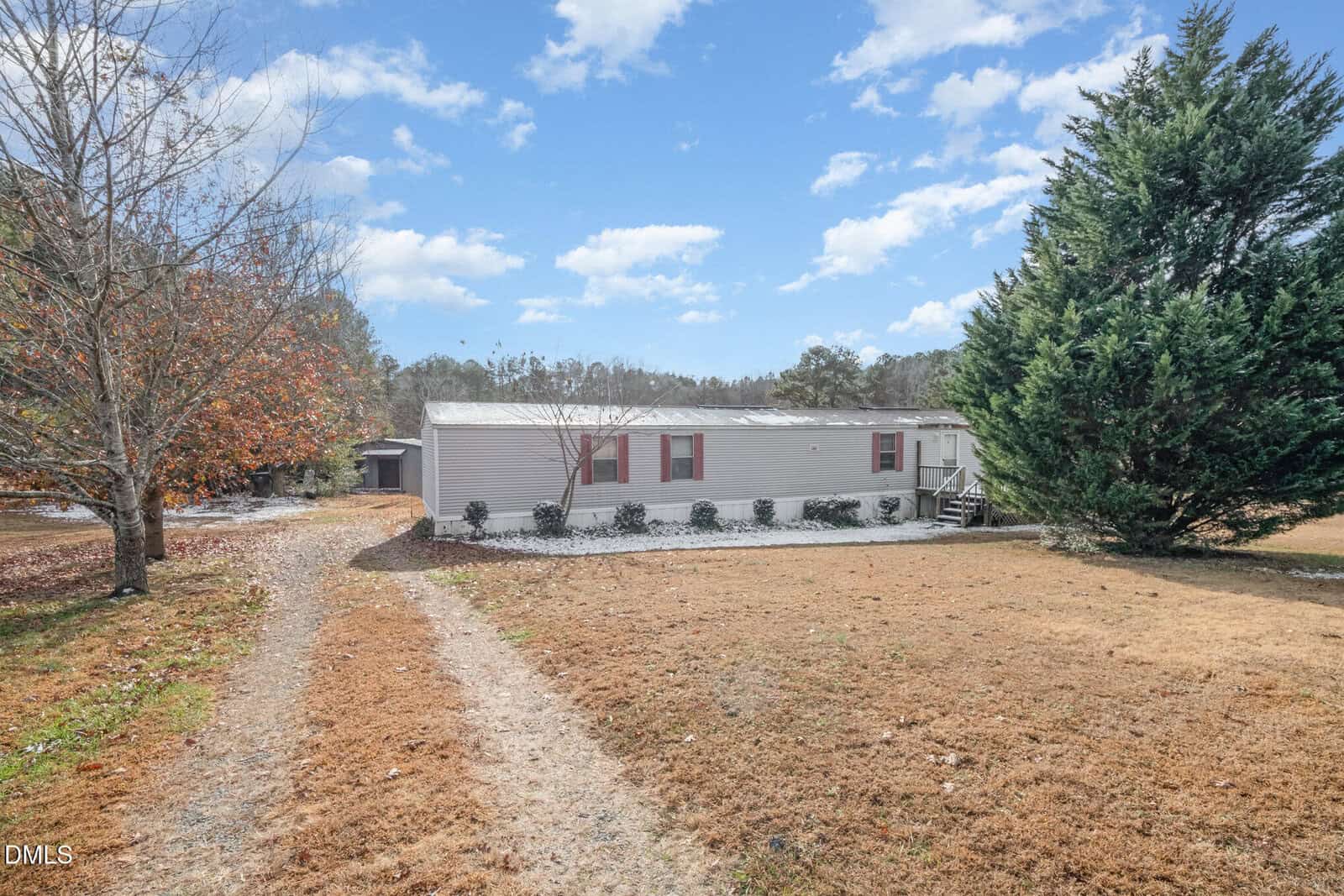 A single-story manufactured home with light-colored siding sits on a grassy lot. There is a dirt driveway leading to the home, a large evergreen tree to the right, and a bare tree to the left. The sky is partly cloudy.