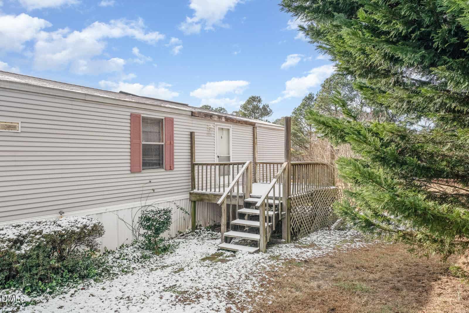 A beige mobile home with red shutters and a small wooden porch with steps and a ramp. Sparse snow covers the ground and bushes, with evergreen trees on the right and a blue sky overhead.