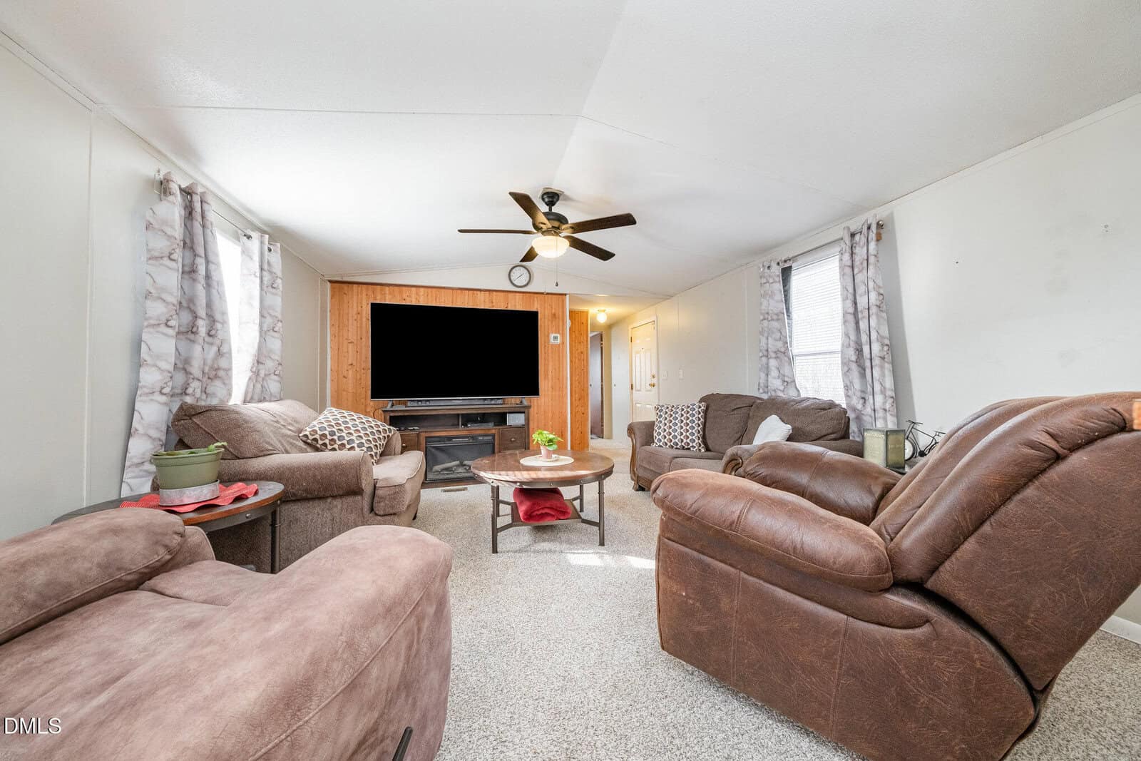 A living room with neutral walls, carpeted floor, brown upholstered sofas and armchairs, a round coffee table, a TV on a wooden stand, a ceiling fan, and two windows with patterned curtains.